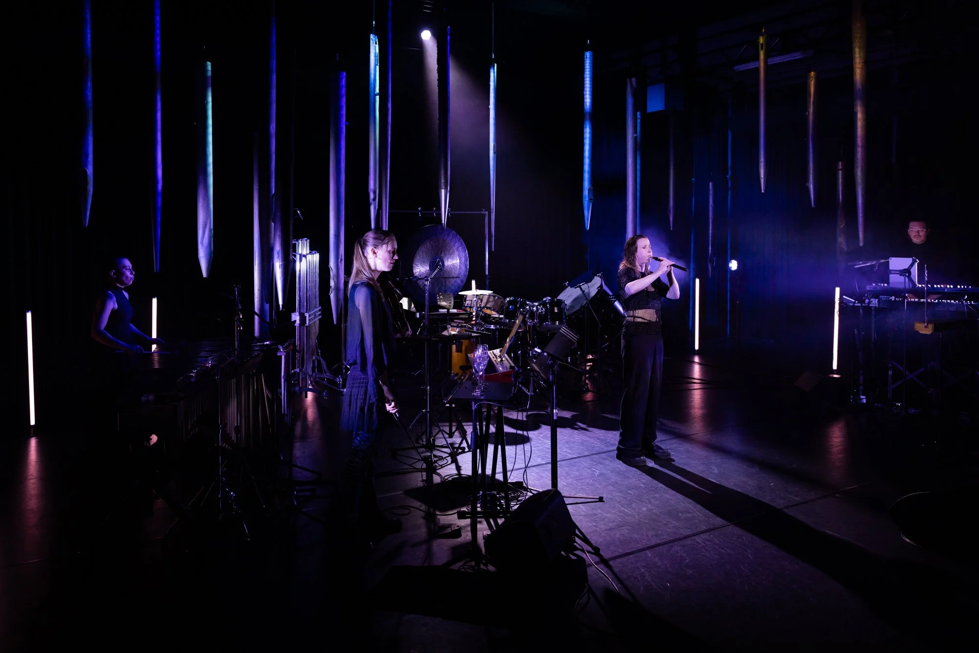 A live music performance on stage with four musicians playing instruments and singing under purple and blue stage lighting, with hanging metallic ornaments in the background.