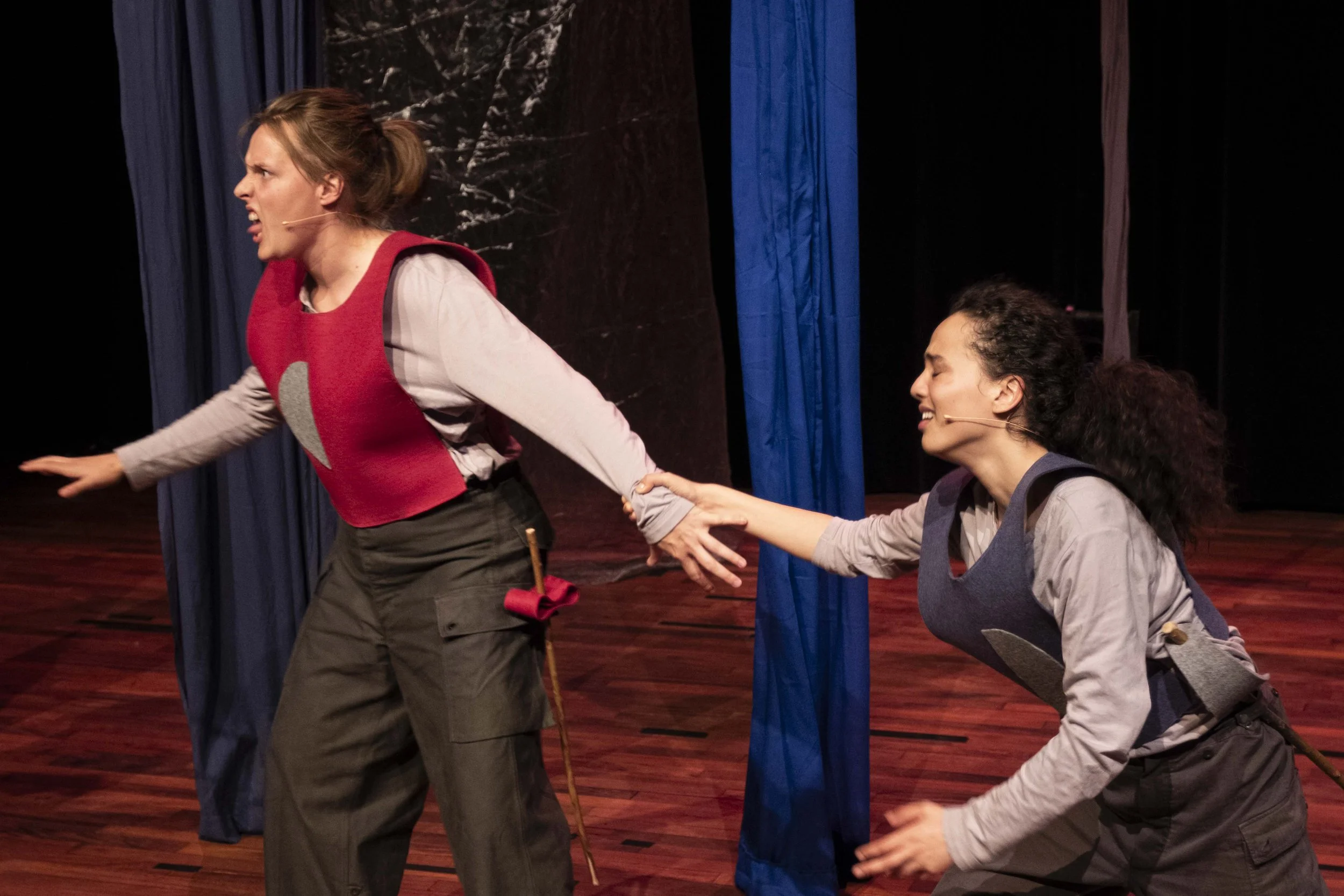 Two women performing on stage, one woman is pulling the other by her arm, both are wearing microphones and colorful vests, with blue curtains in the background.