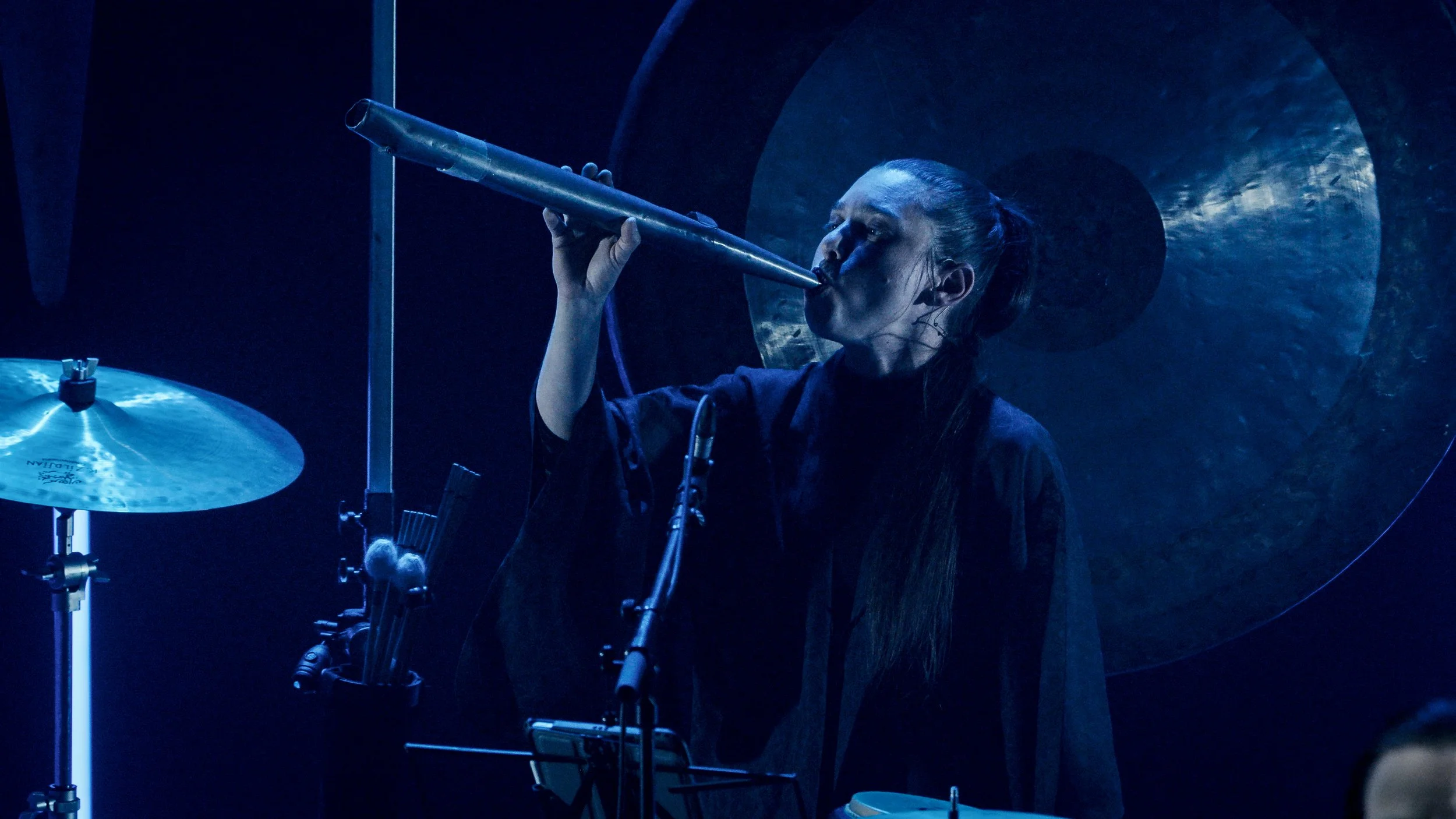 A woman playing a didgeridoo in a dark blue-lit room with a gong in the background.