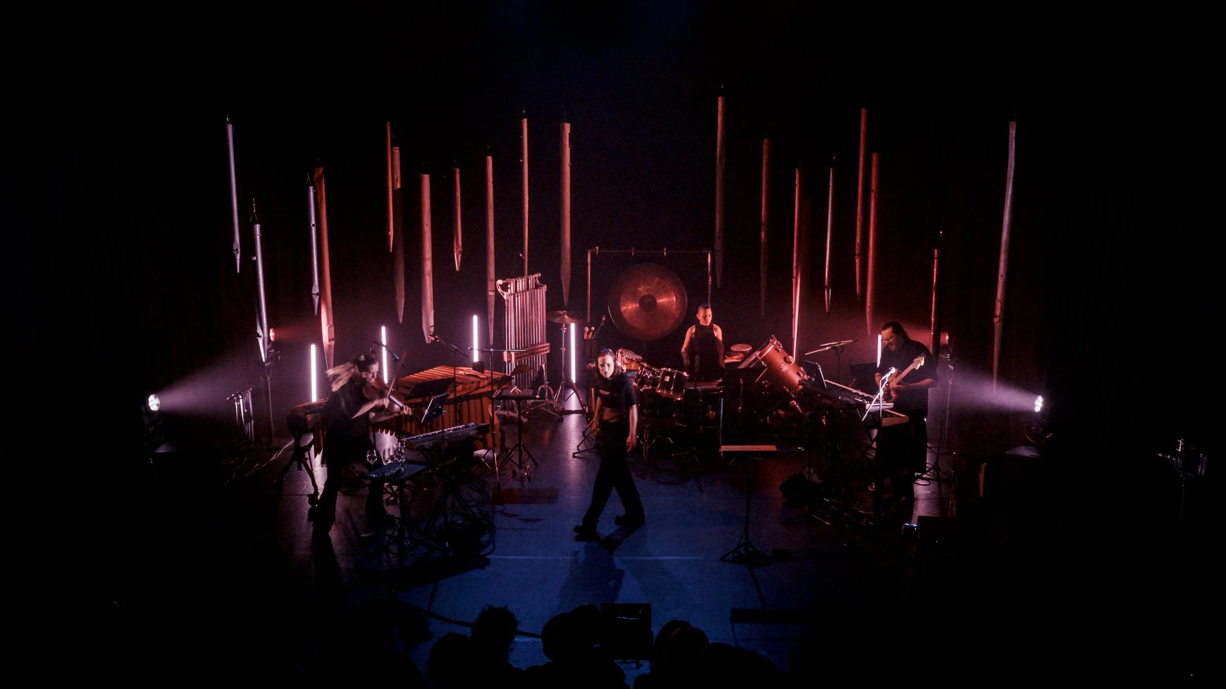 A band performing on stage with various percussion instruments, illuminated by pink and purple stage lights, with an audience in front.