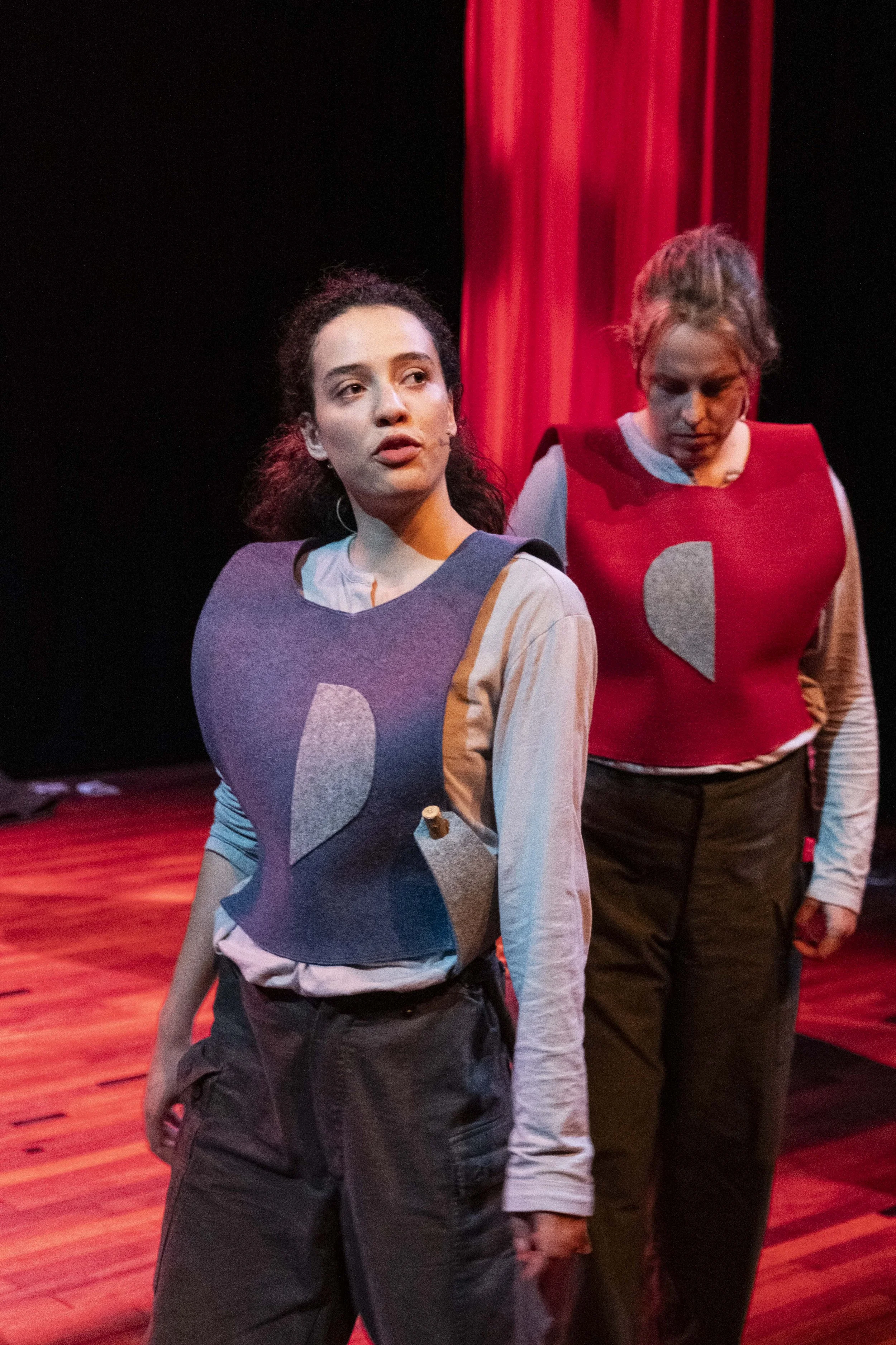 Two women on stage wearing felt breastplates with broken heart symbols, performing in a theatrical production