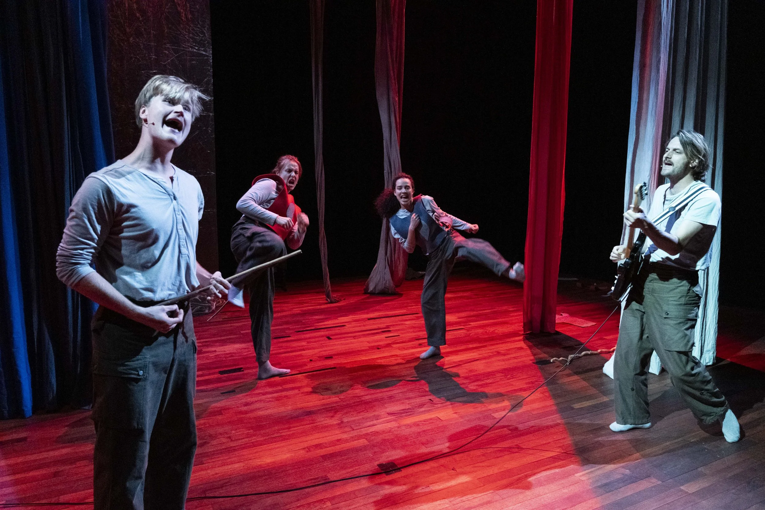 Four actors performing on stage with red and blue curtains, illuminated by stage lights, engaged in energetic acting with musical and theatrical expressions.