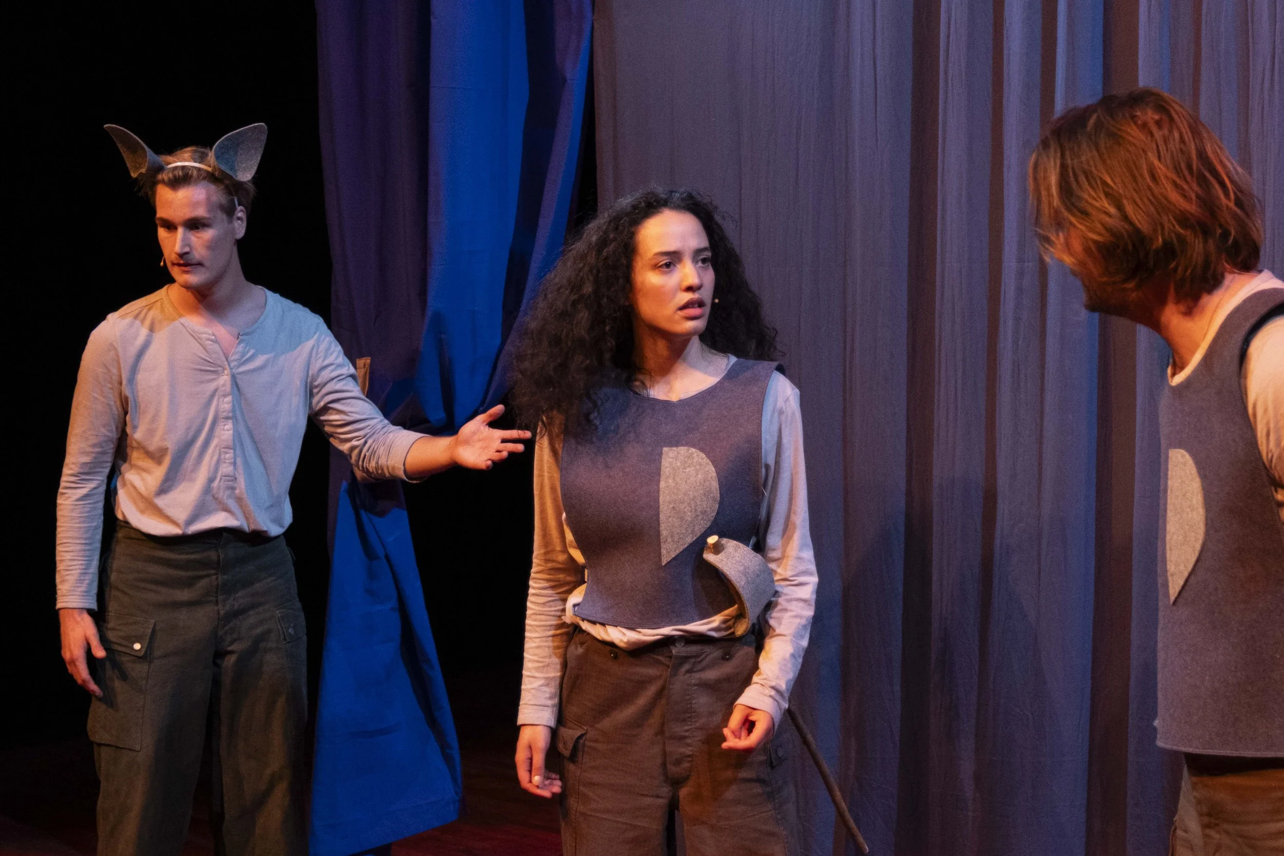 Three actors on stage; one with fox ears costume, one woman with dark curly hair and a vest, and another with long hair, all engaged in a scene.