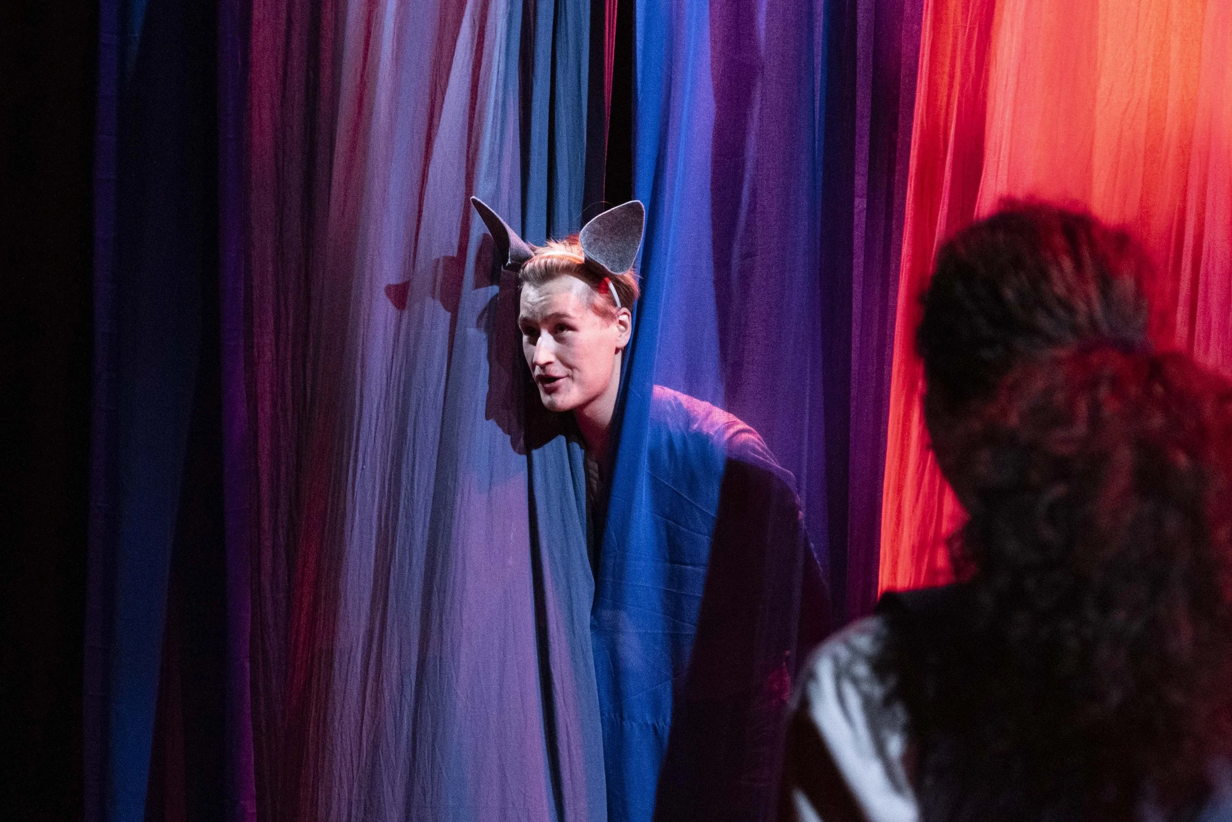 A person dressed as a mouse with gray ears and red hair peeks through colorful theater curtains while talking to a woman with curly hair.