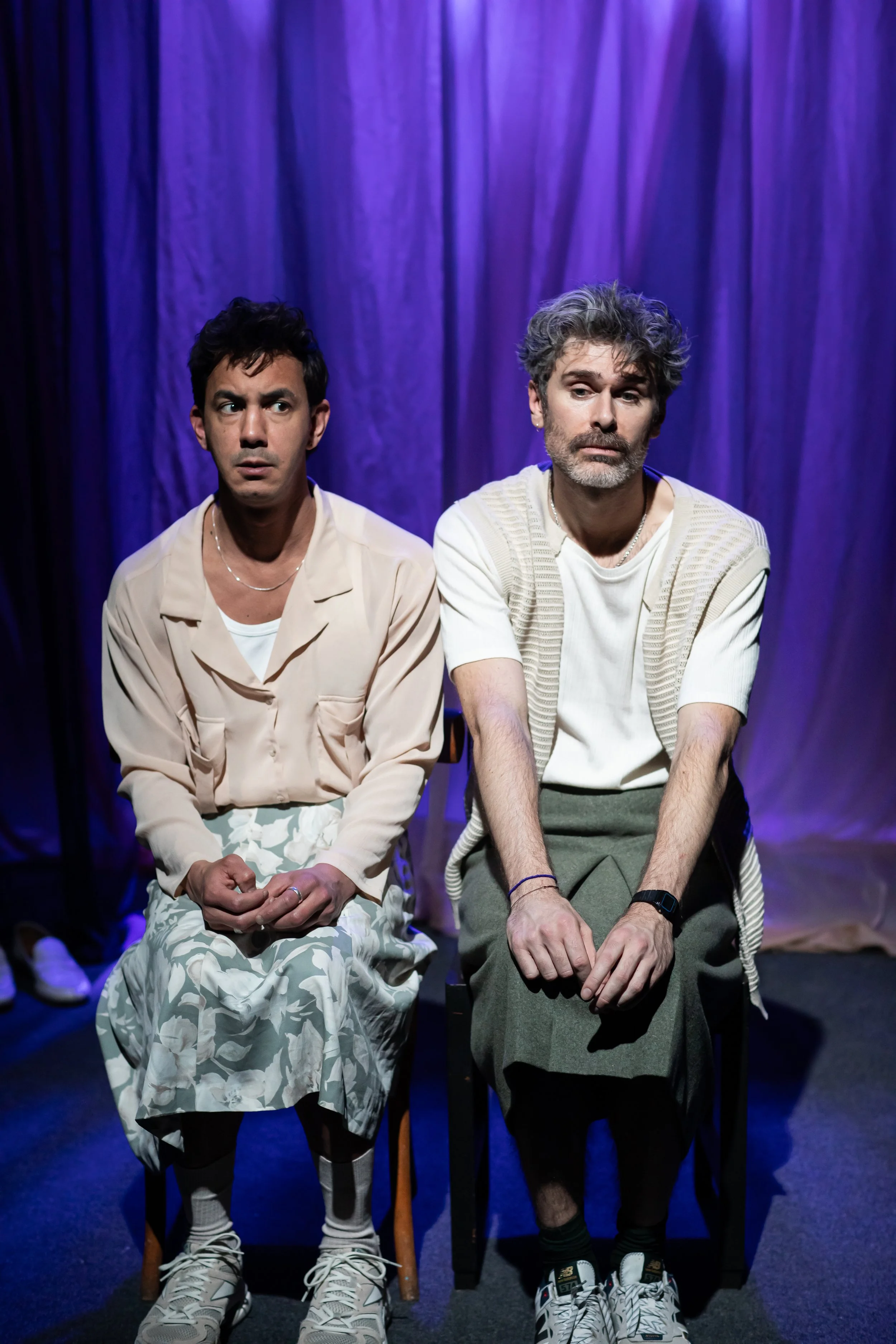 Two men sitting on chairs in front of purple curtains, appearing to be in a theatrical performance.