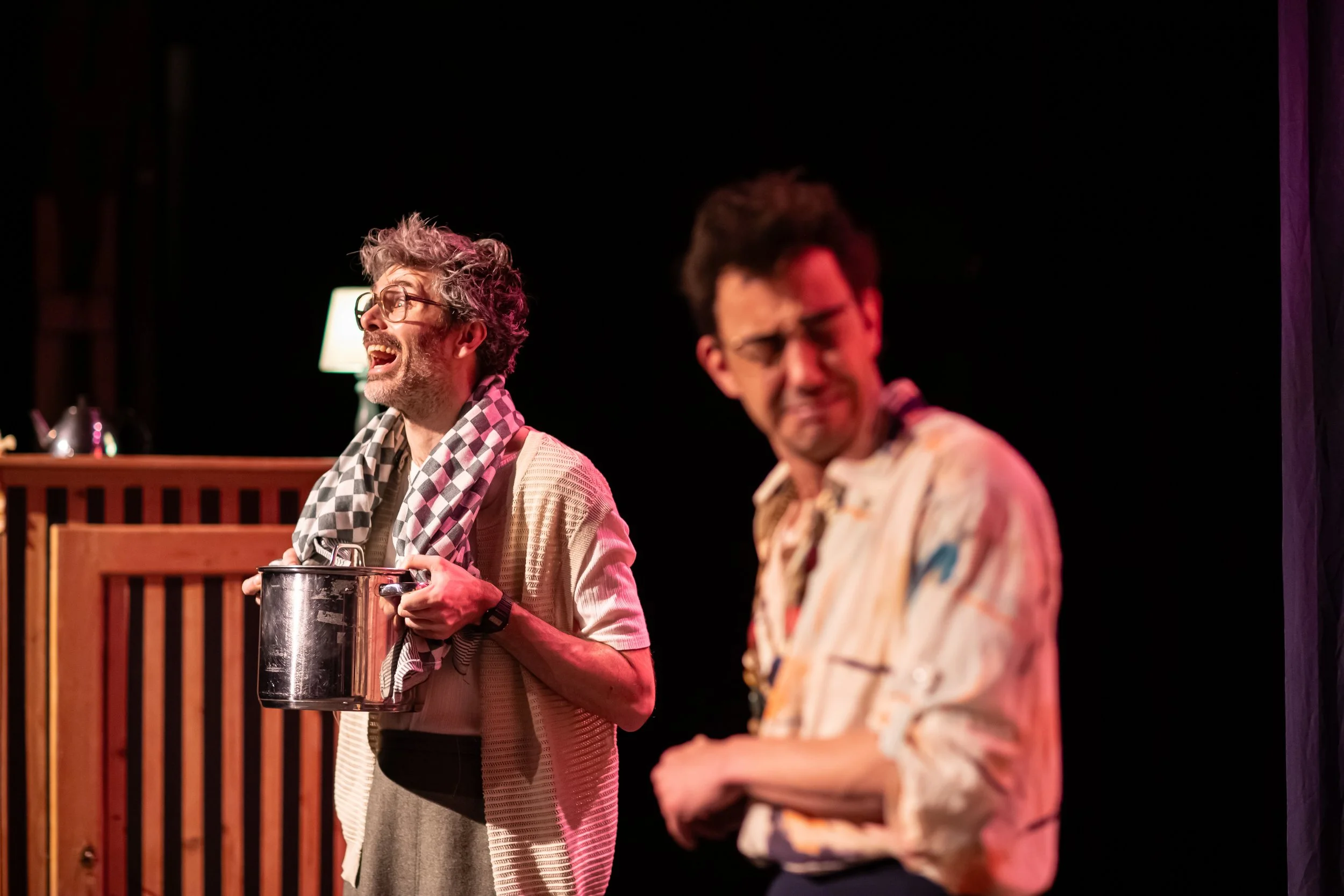 Two men acting on stage, one holding a pot and laughing, the other looking confused or upset, with a dark background.