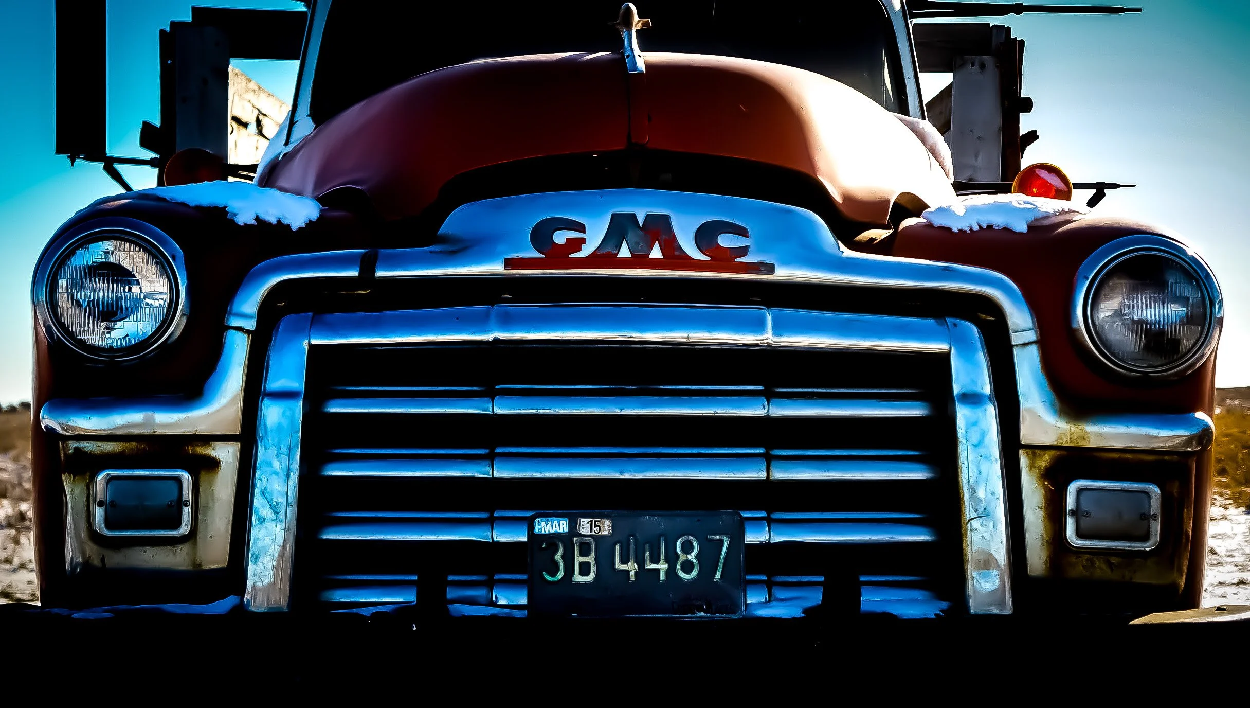 Close-up front view of a vintage red GMC truck with license plate 3B4487, snow on the fenders, and a clear blue sky background.