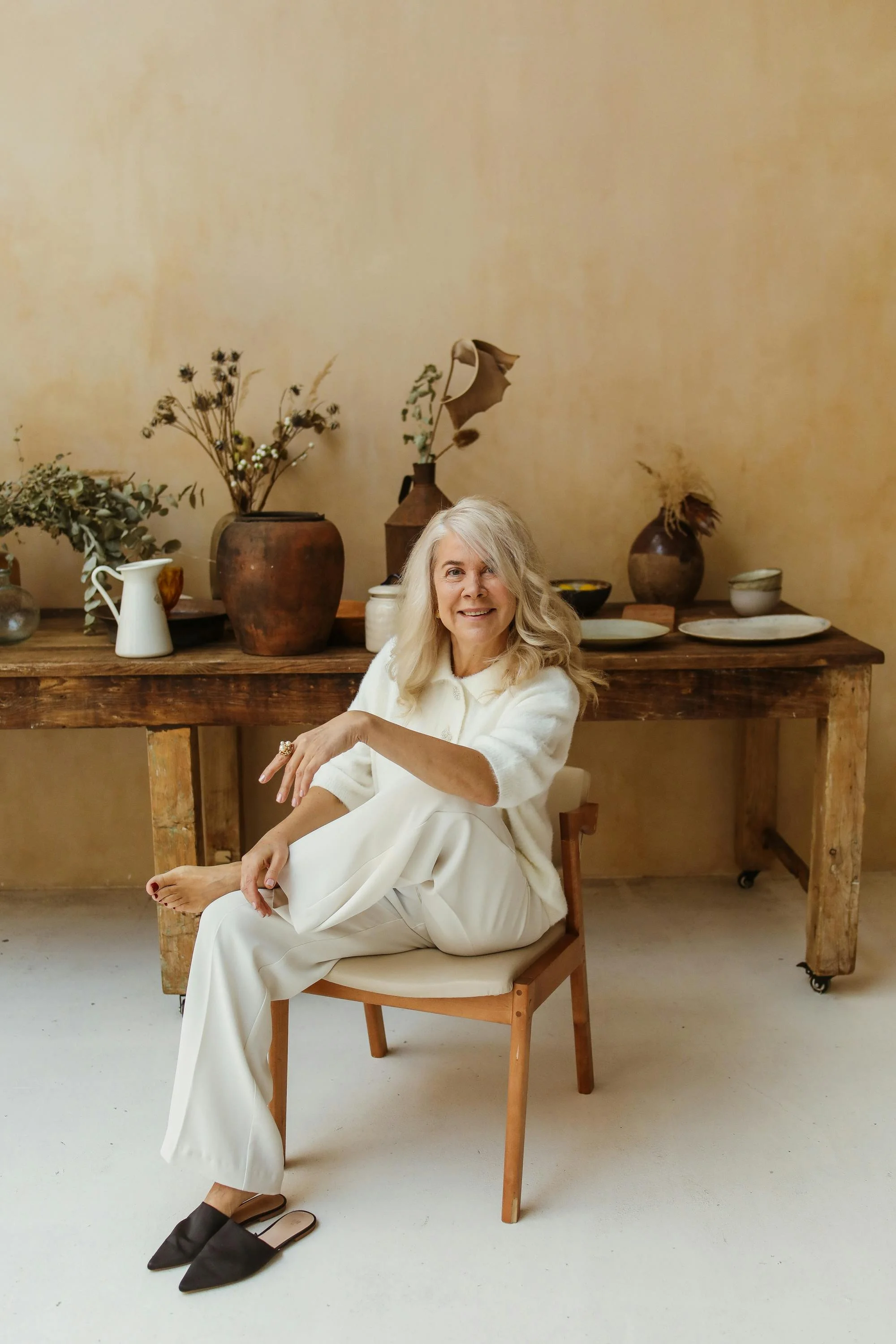 A smiling elderly woman with long gray hair sits on a wooden chair in front of a rustic wooden table with decorative pottery and dried flowers, against a beige wall.