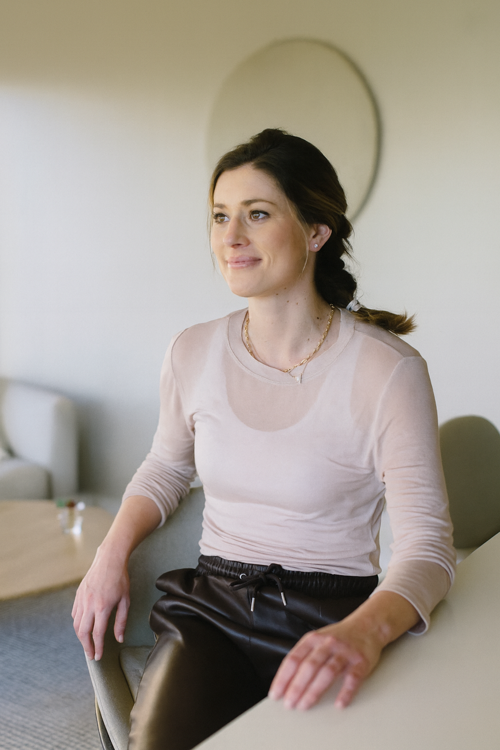 A woman with brown hair tied in a braid, wearing a light beige long-sleeve top and black drawstring pants, sitting at a table in a modern, well-lit room.