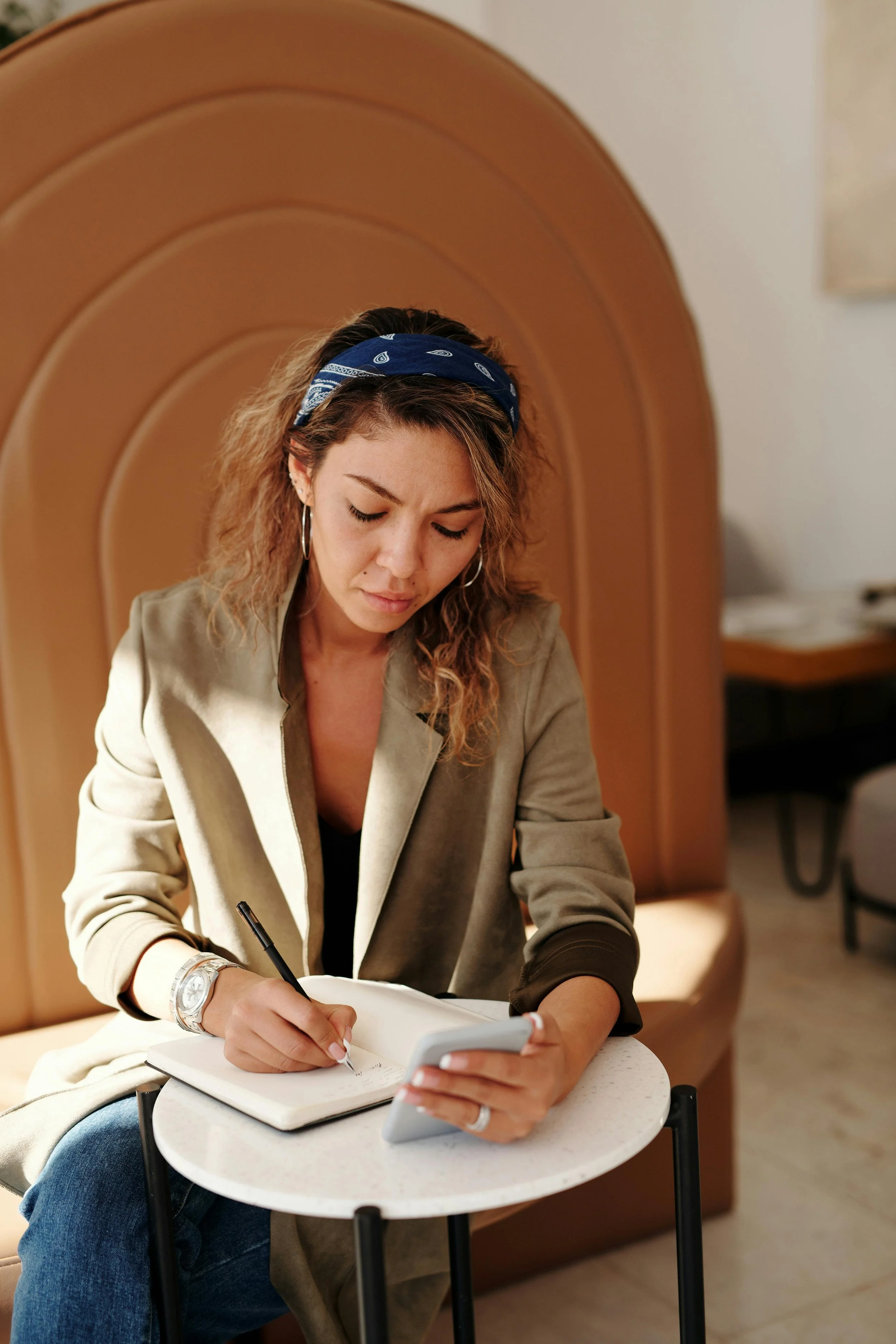 A woman with curly hair wearing a blue bandana, earrings, and a beige jacket sitting at a small round table, writing in a notebook and looking at a smartphone.