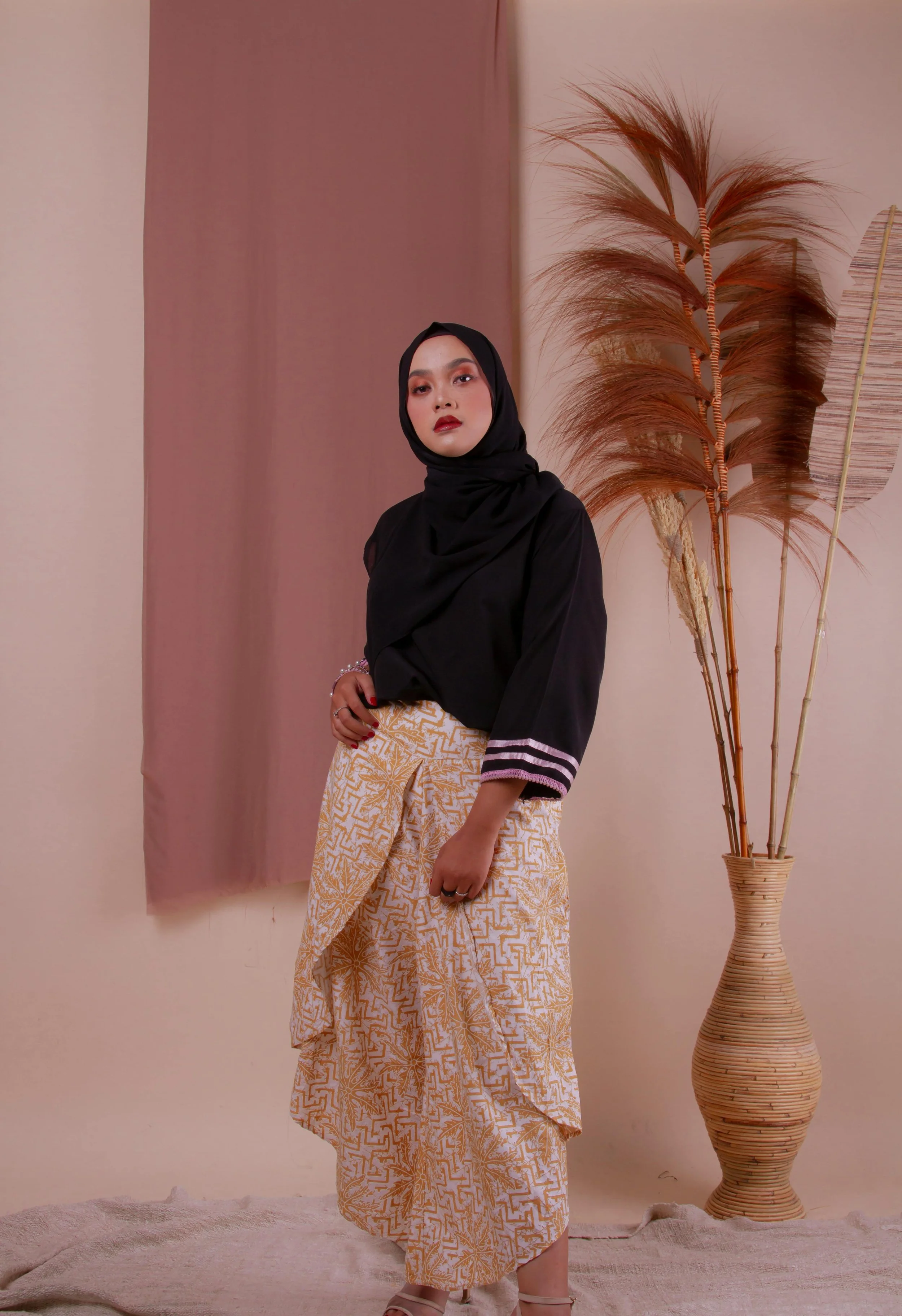 A woman wearing a black hijab and black top with striped sleeves stands indoors next to a large decorative vase with dried plants. The background includes draped fabric and a peach-colored wall.