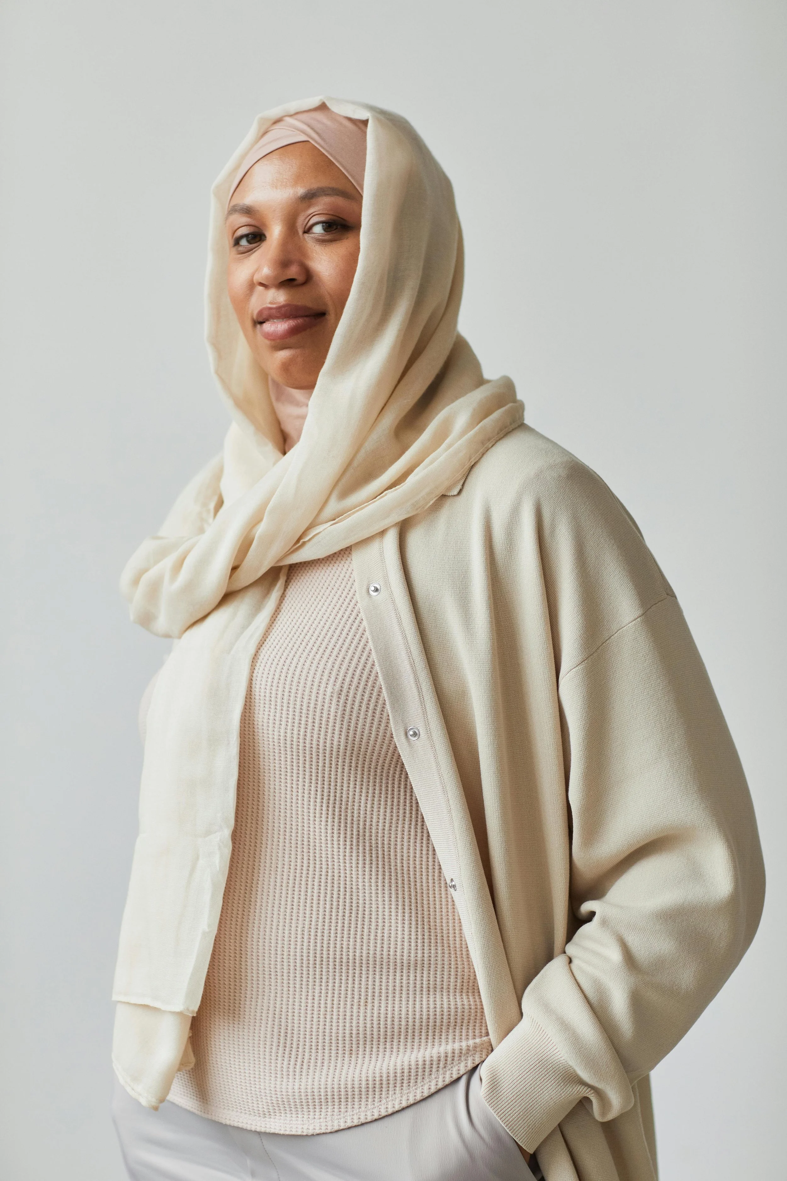 A woman wearing a beige hijab, cream-colored hoodie, and a beige textured top, standing against a plain light background.