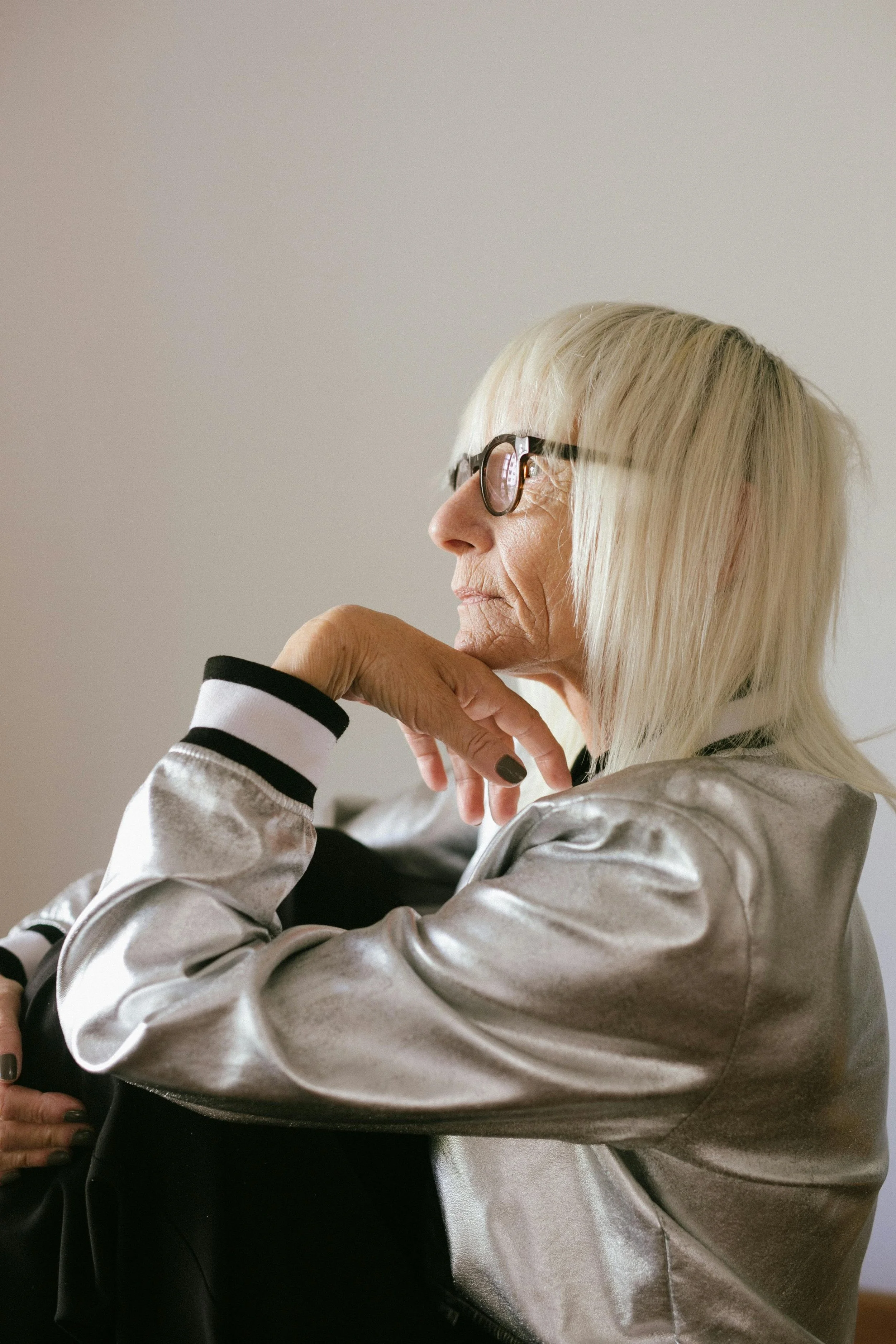 Profile of elderly woman with blonde hair wearing glasses and a silver jacket, sitting with her hand resting on her chin, looking thoughtful.