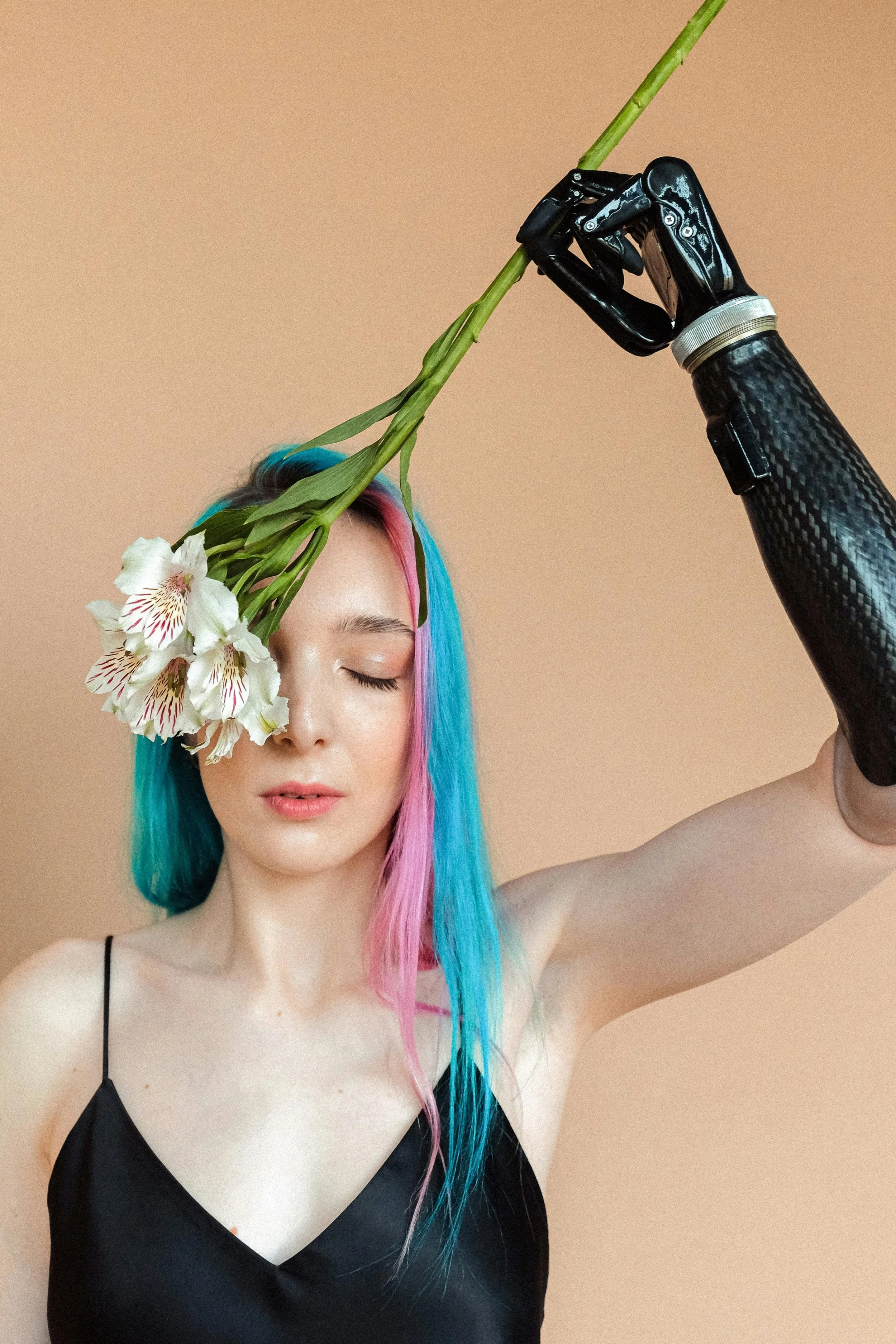 A woman with pastel-colored hair holding a flower near her forehead while her hand, which appears robotic, holds the stem of the flower.