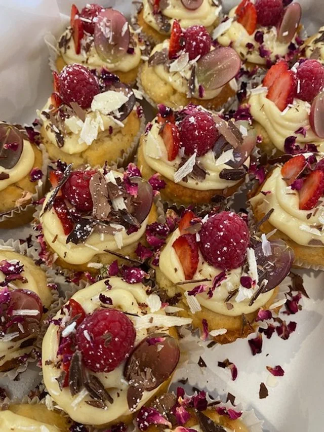 Mini cupcakes topped with white frosting, fresh raspberries, sliced strawberries, chocolate shavings, and edible flower petals, decorated with sprinkles.