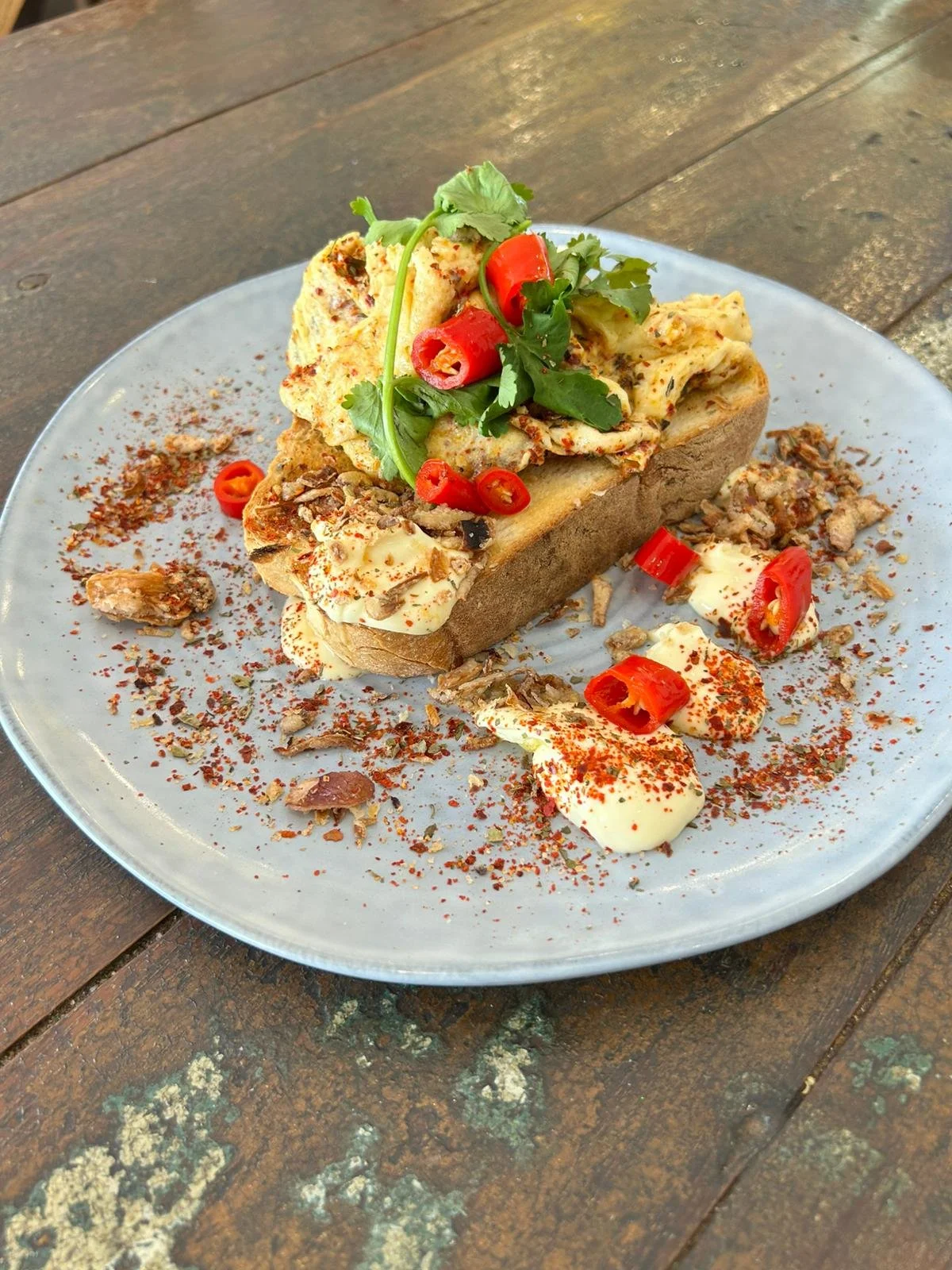 A plate of loaded French bread with various toppings including whipped cream, chopped chili peppers, herbs, and seasoning, garnished with cilantro and red chili slices, on a wooden table.