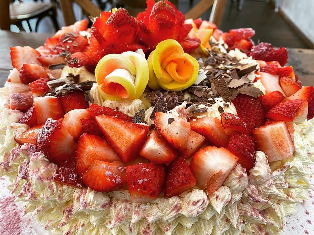 A large decorated fruit cake topped with fresh strawberries, sliced strawberries, chocolate shavings, and decorative roses made of marzipan or icing.