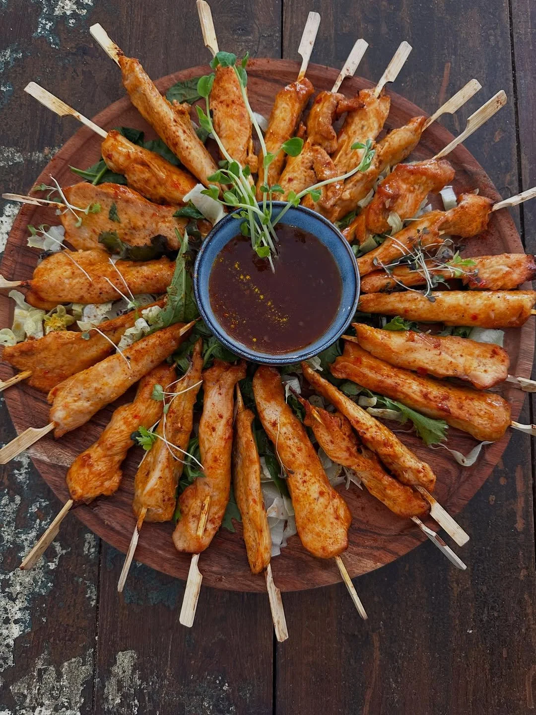 A round wooden platter filled with grilled chicken skewers arranged in a circle, with fresh greens and microgreens in the center, and a small bowl of dipping sauce in the middle.