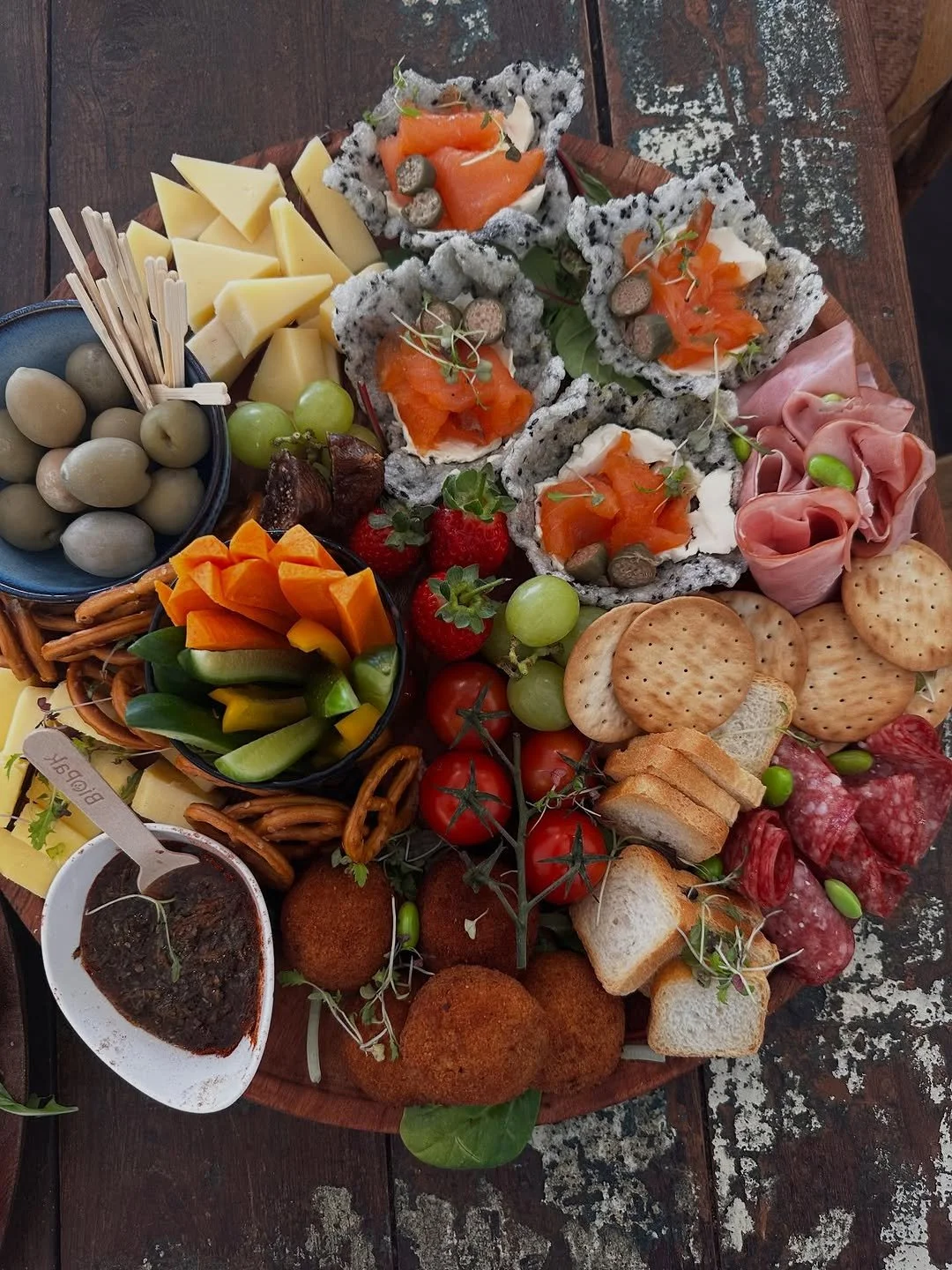A large cheese and charcuterie platter with crackers, vegetables, fruit, olives, smoked salmon, and various cheeses on a wooden table.