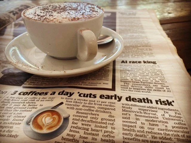 A white cup filled with a frothy coffee, sitting on a matching saucer with a spoon. The cup and saucer are placed on a newspaper on a wooden table.