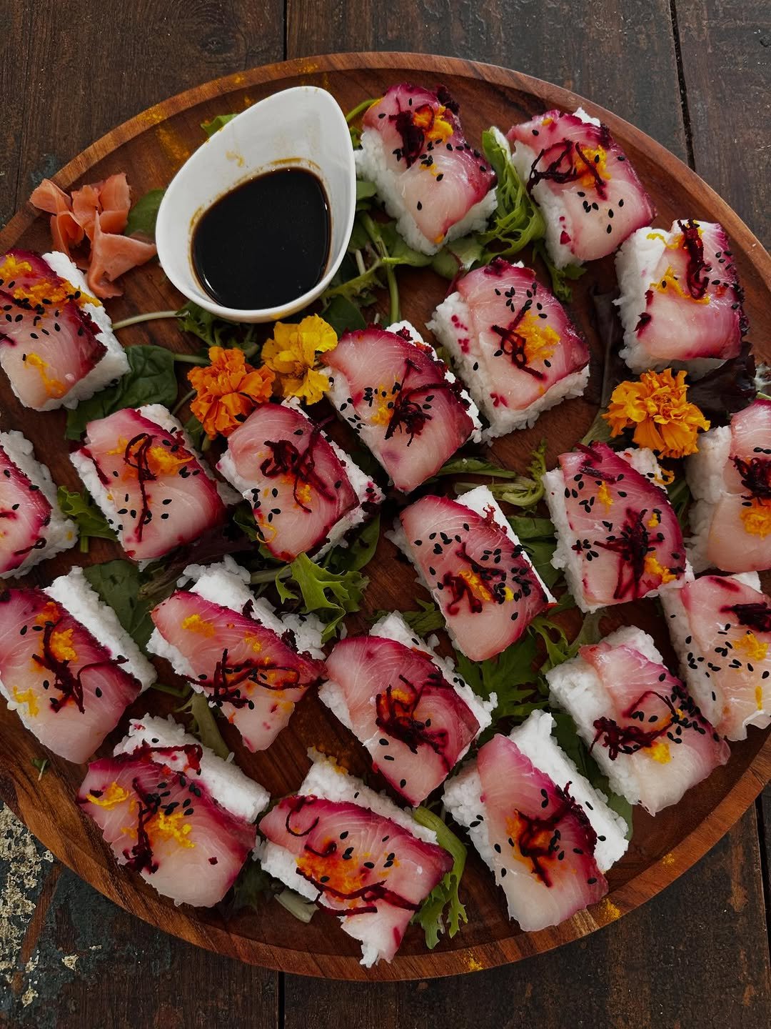 A round wooden serving tray with multiple pieces of sushi topped with fish and garnished with colorful sauces, black sesame seeds, and edible flowers, along with a small dish of soy sauce in the center.