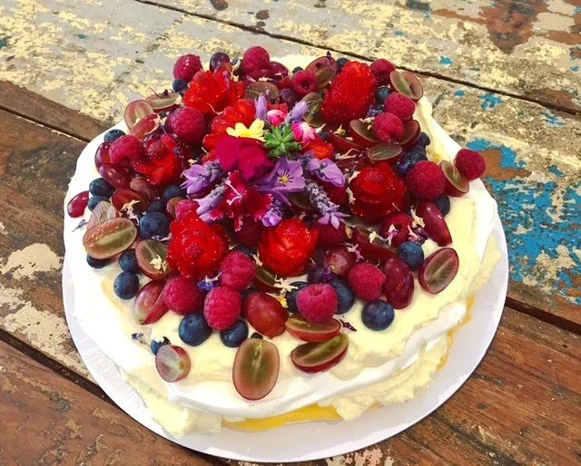 A cake topped with mixed berries including raspberries, blueberries, red currants, and halved grapes, decorated with purple flowers and placed on a white plate on a rustic wooden table.