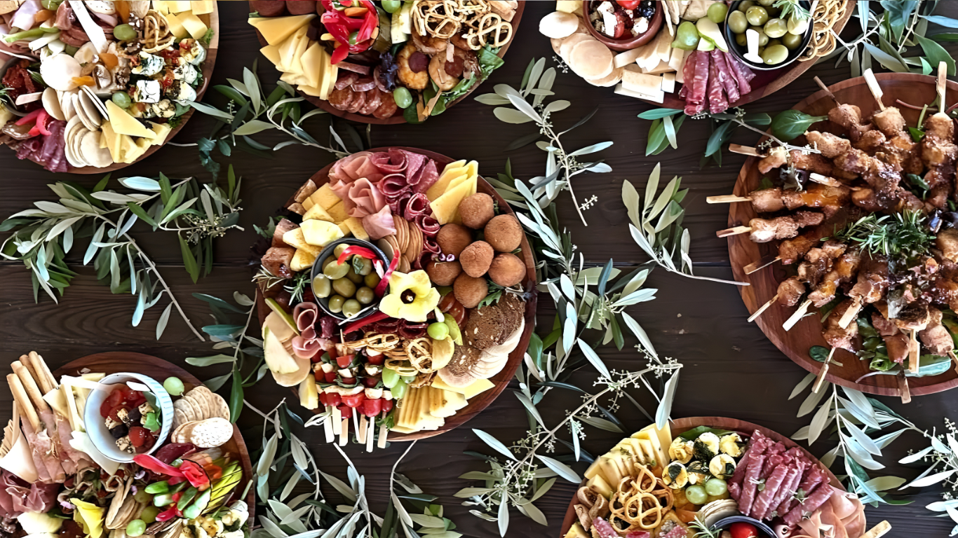 Assorted charcuterie and appetizer platters with meats, cheeses, fruits, vegetables, and crackers, displayed on a dark wooden table decorated with greenery.
