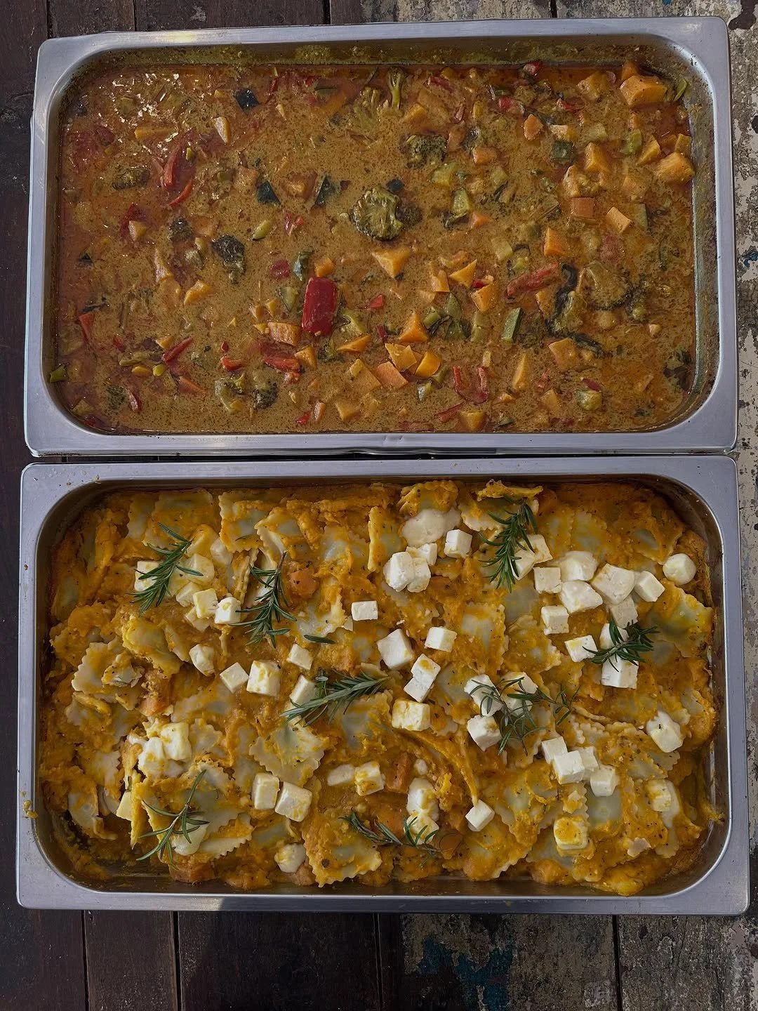 Two stainless steel trays of cooked food, the top tray contains a vegetable stew with chopped vegetables and sauce, and the bottom tray contains baked pasta with cheese, garnished with sprigs of rosemary and small cubes of cheese.