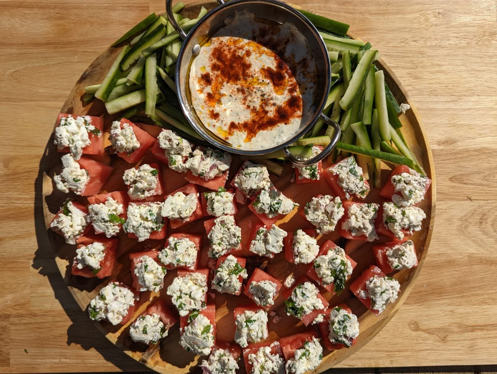 A round wooden platter with cubed watermelon topped with crumbled feta cheese, sliced cucumbers, and a small pot of creamy dip garnished with paprika.