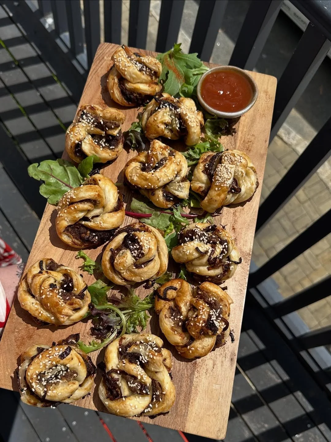 A wooden serving tray with multiple savory pastries topped with sesame seeds and drizzled with chocolate, garnished with leafy greens, and accompanied by a small bowl of ketchup or dipping sauce, set outdoors on a balcony.