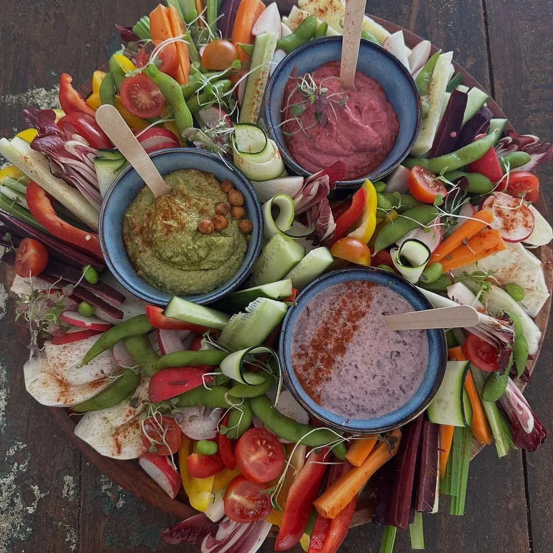 Colorful vegetable platter with cherry tomatoes, carrots, cucumbers, radishes, bell peppers, and microgreens, served with three bowls of different dips or spreads.