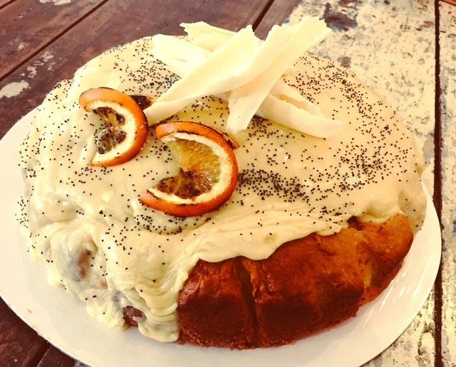 Plate of a dessert with whipped cream, orange slices, white chocolate shards, poppy seeds, and a cake with caramelized edges on a rustic wooden table.