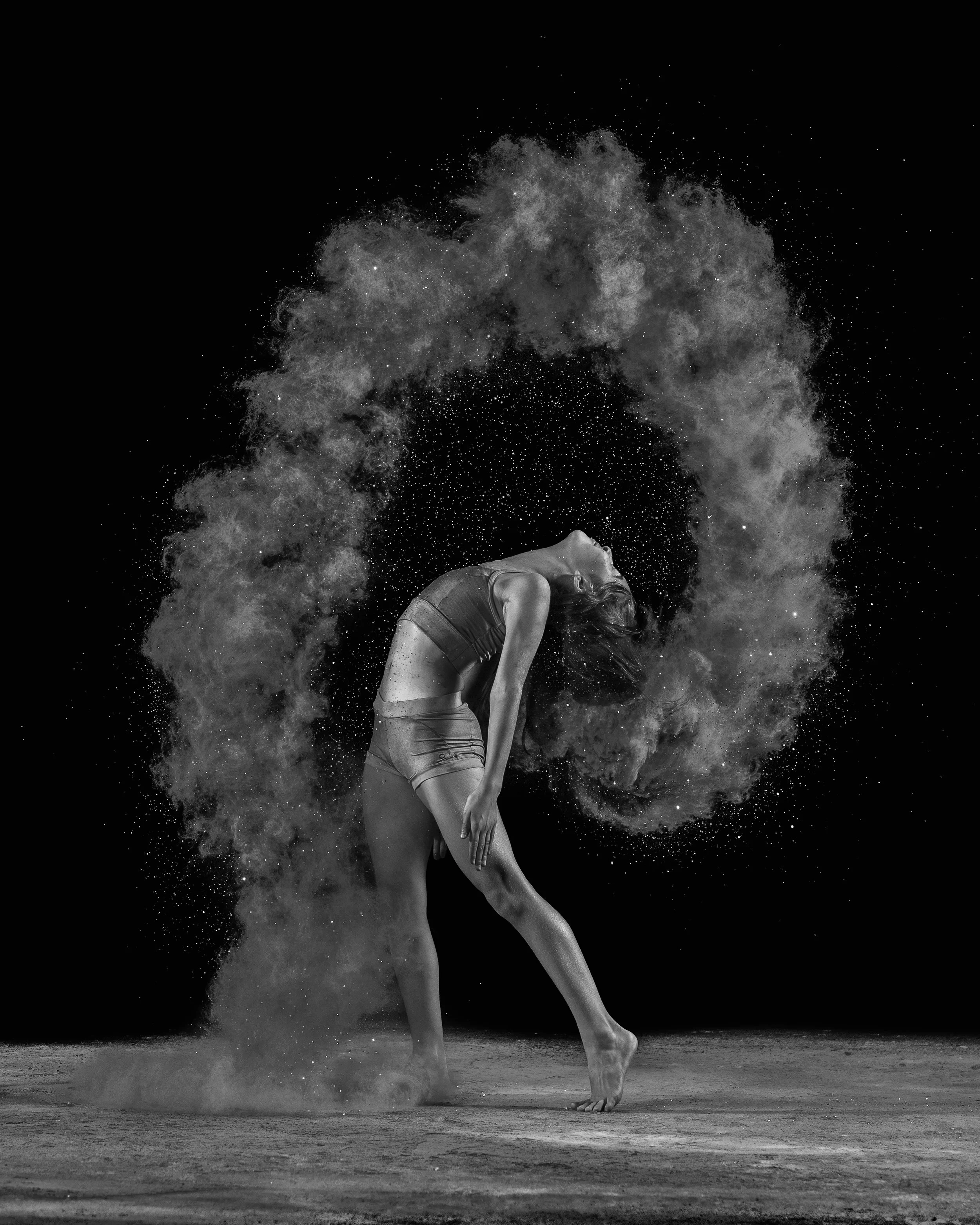 A woman in athletic attire performing a dance move surrounded by a cloud of powdered substance against a black background.