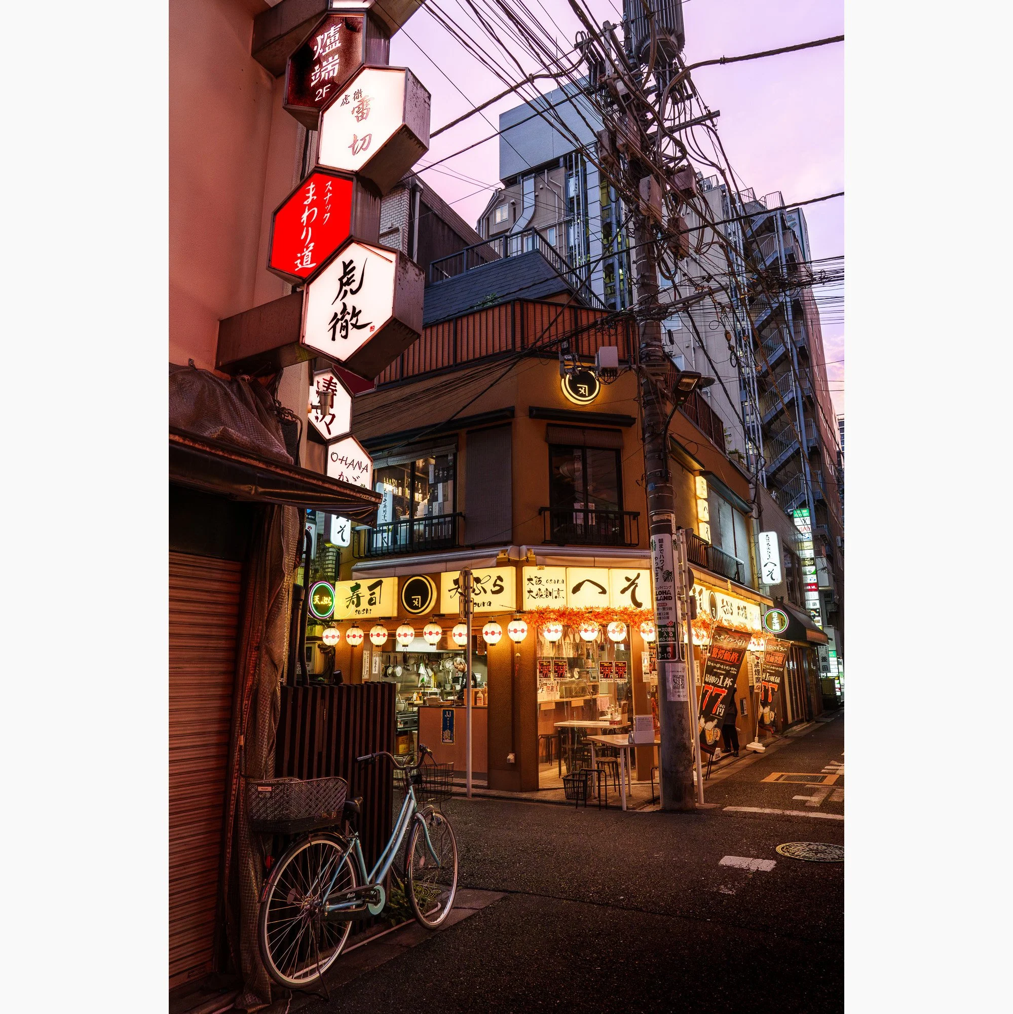 Evening Tokyo street scene with illuminated Japanese signs, tangled overhead power lines, bicycles and glowing restaurants at dusk.