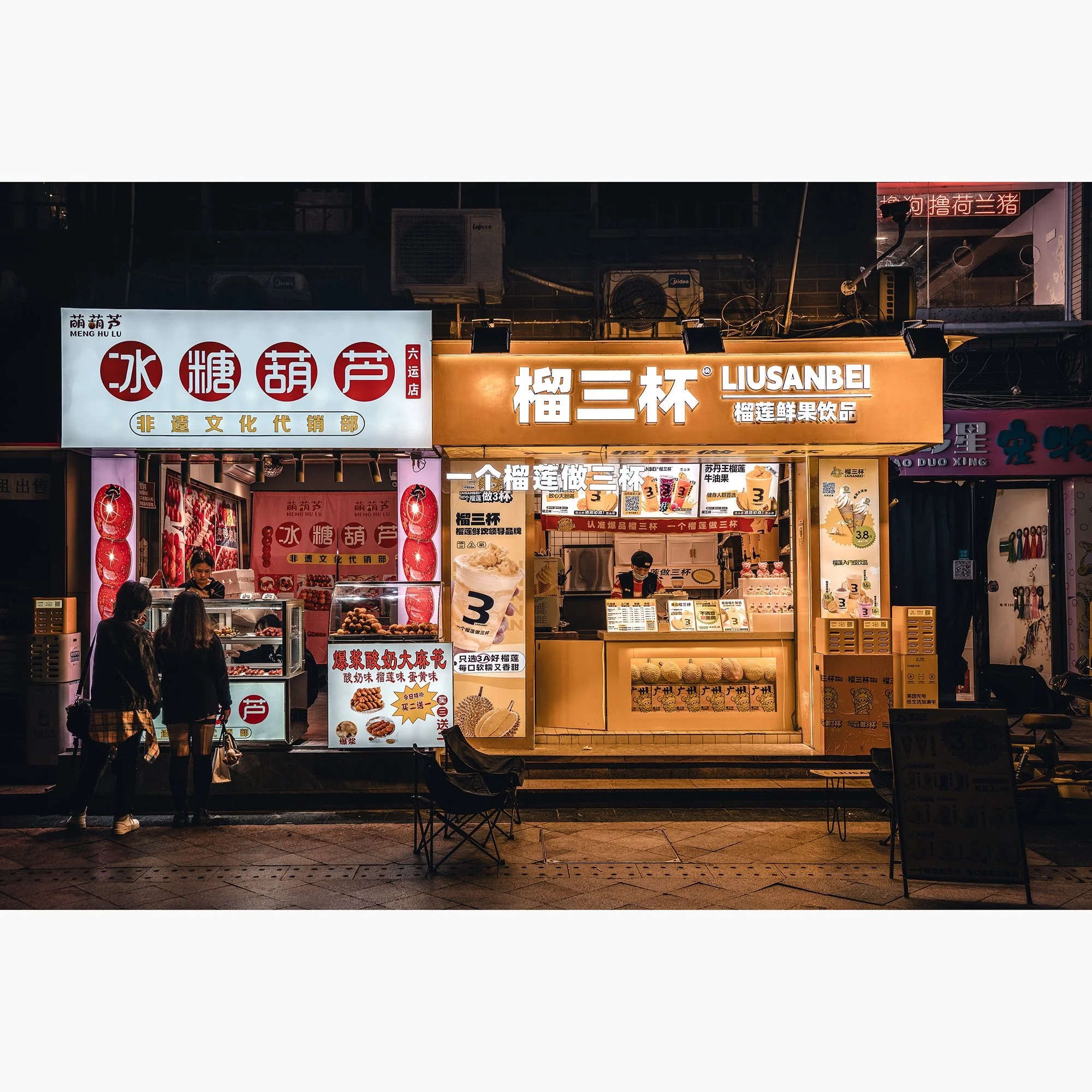 Night street scene in Guangzhou, China with illuminated food stalls, Chinese signage, warm orange lighting and pedestrians in front of small storefronts.