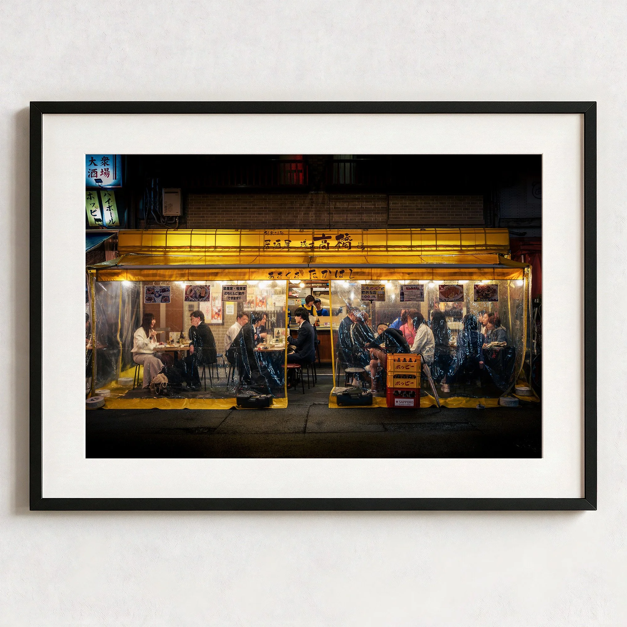 Framed Tokyo night street photography print showing a busy Japanese izakaya beneath a glowing yellow awning, displayed in a black frame with white mount.