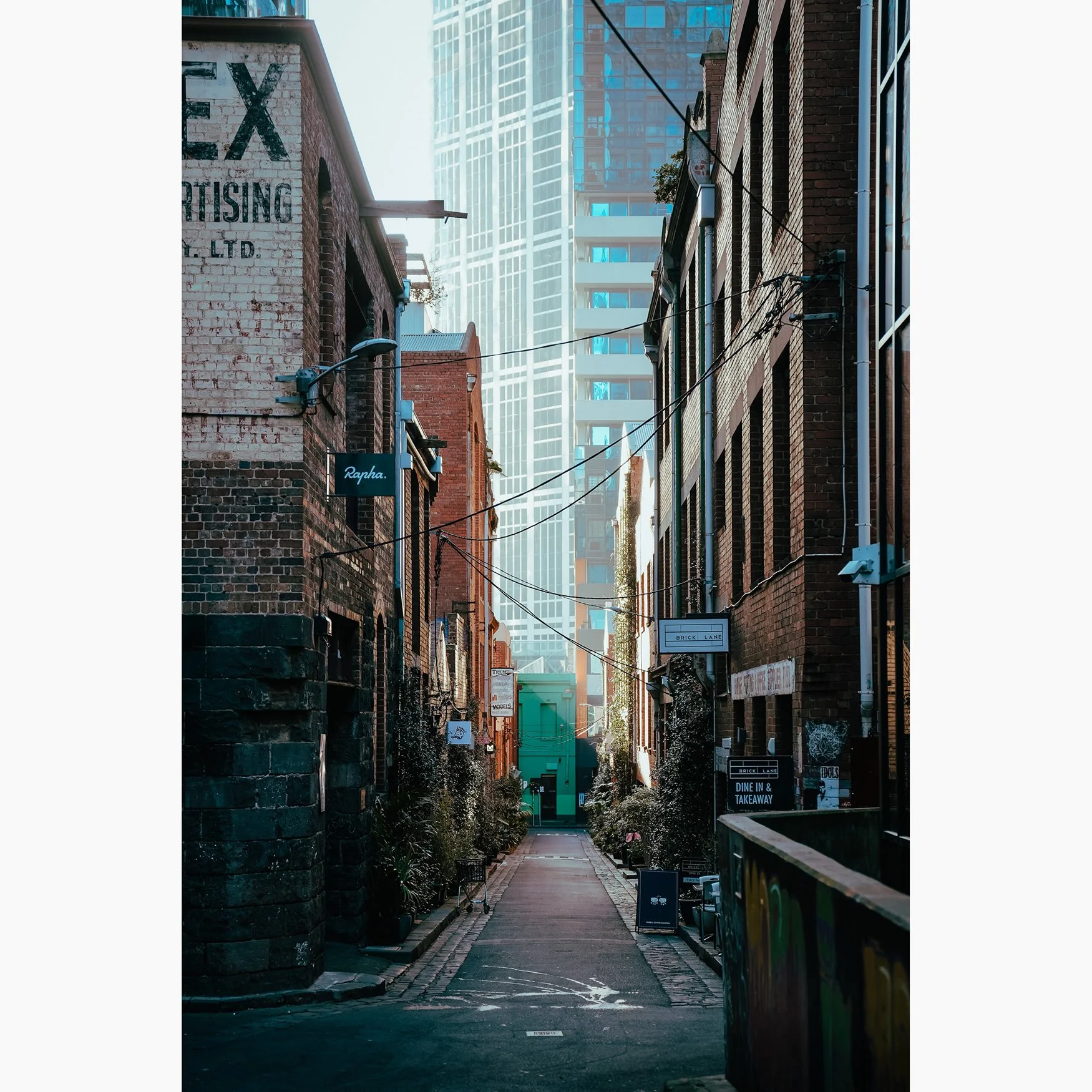 Vertical urban photograph of a narrow Melbourne laneway with brick buildings, overhead wires and modern city towers in the background.