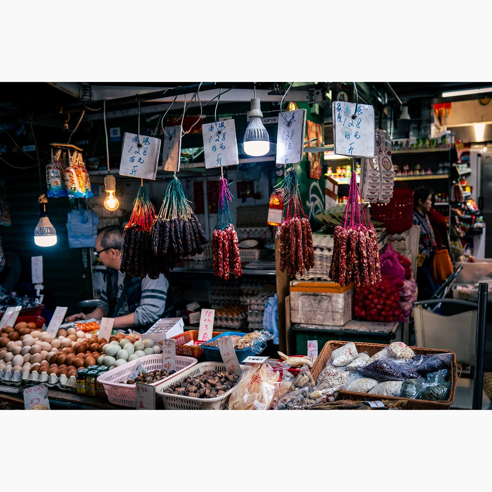 Hong Kong market photograph featuring hanging Chinese sausages, handwritten signs, glowing bulbs, eggs, dried goods and market vendors.