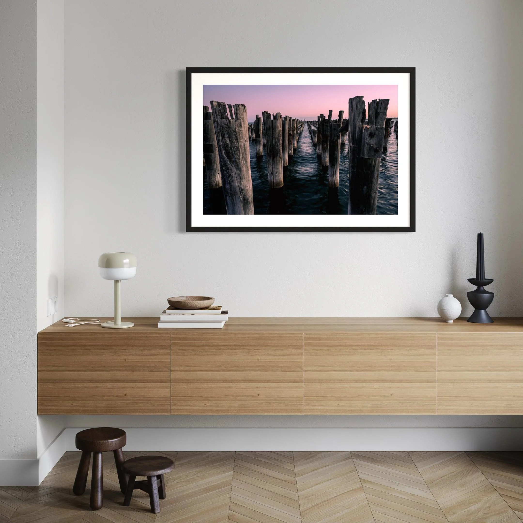 Framed Princes Pier photography print displayed above a timber console in a contemporary interior, showing weathered pylons and soft pink dusk tones.