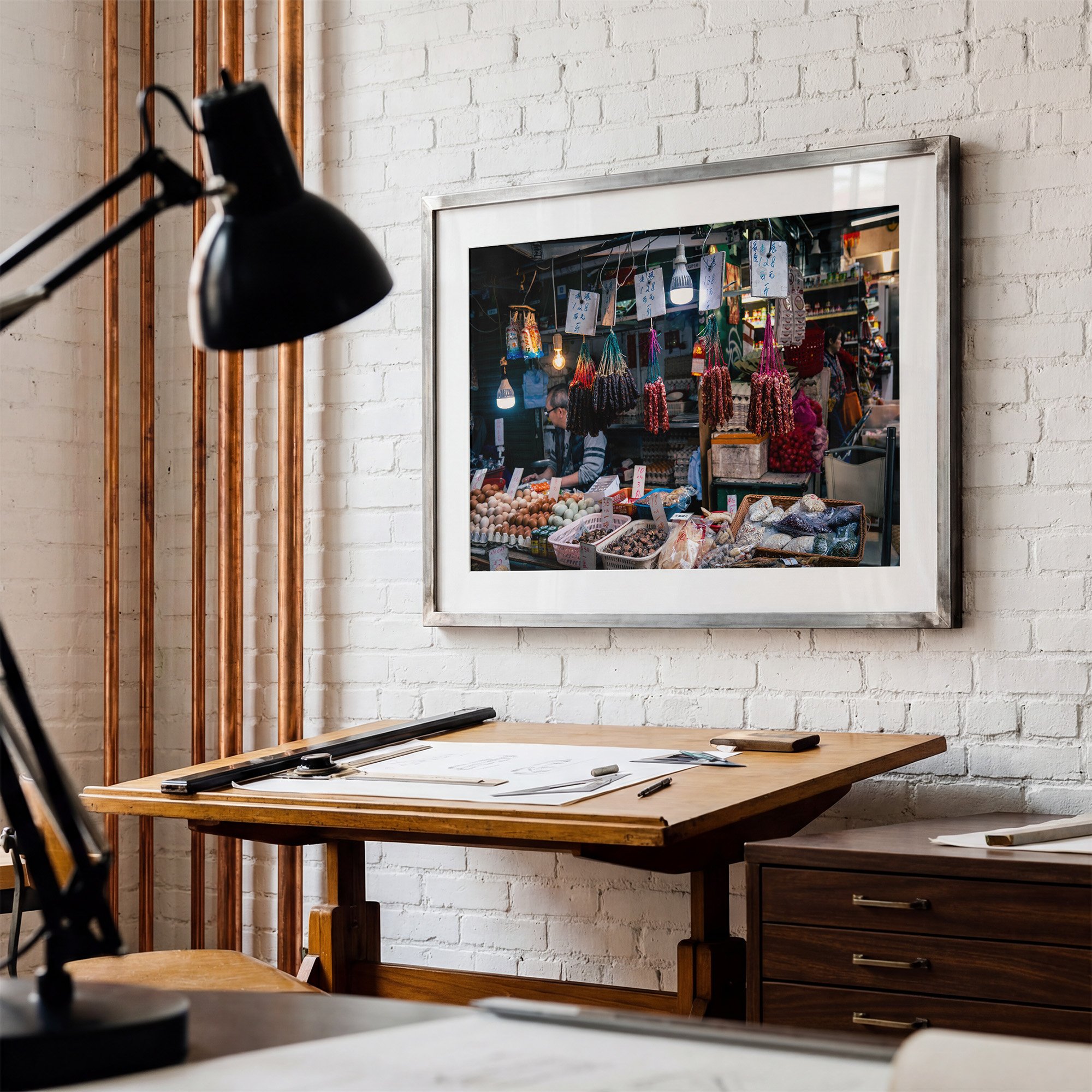 Hong Kong market photography print displayed in a silver frame with white mat on a white brick wall above a wooden drafting table.