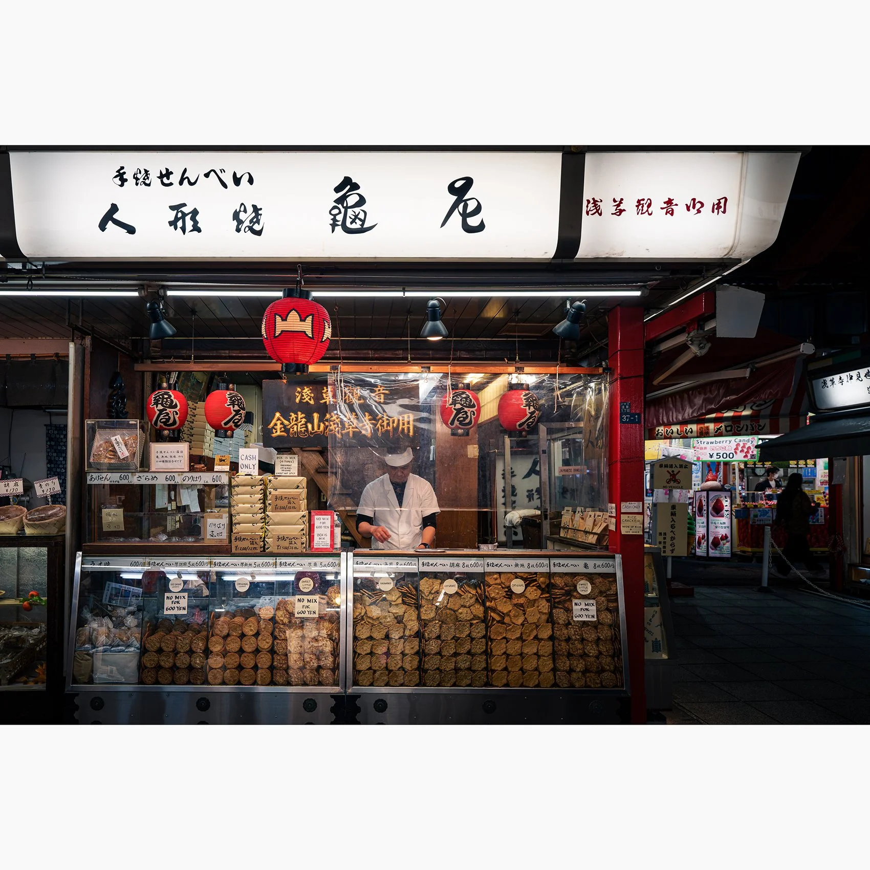 Traditional cracker shop in Asakusa, Tokyo, with a shopkeeper surrounded by senbei displays, lanterns and Japanese signage.