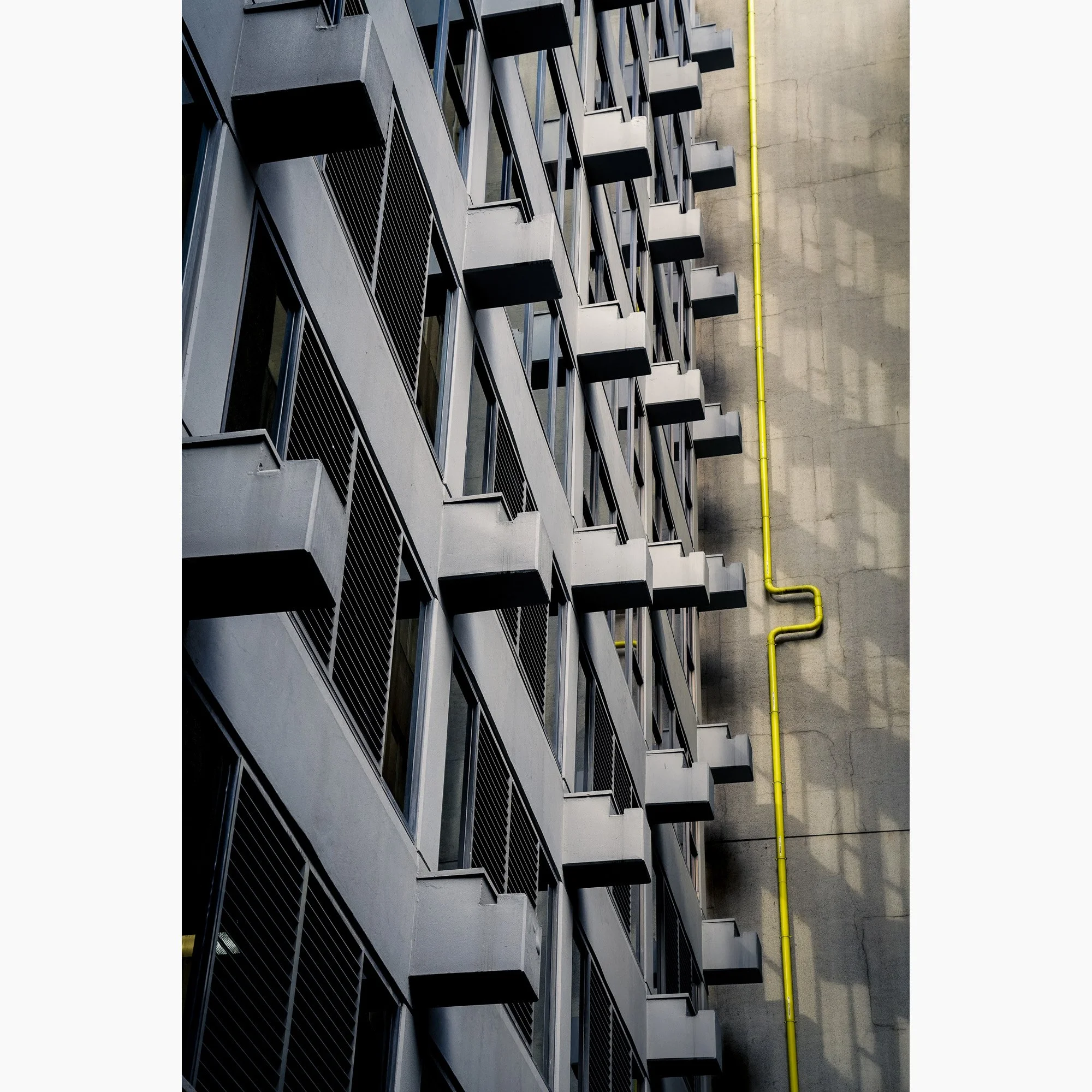 Architectural photograph of repeating concrete balconies and a vivid yellow pipe creating geometric patterns against a textured city wall.