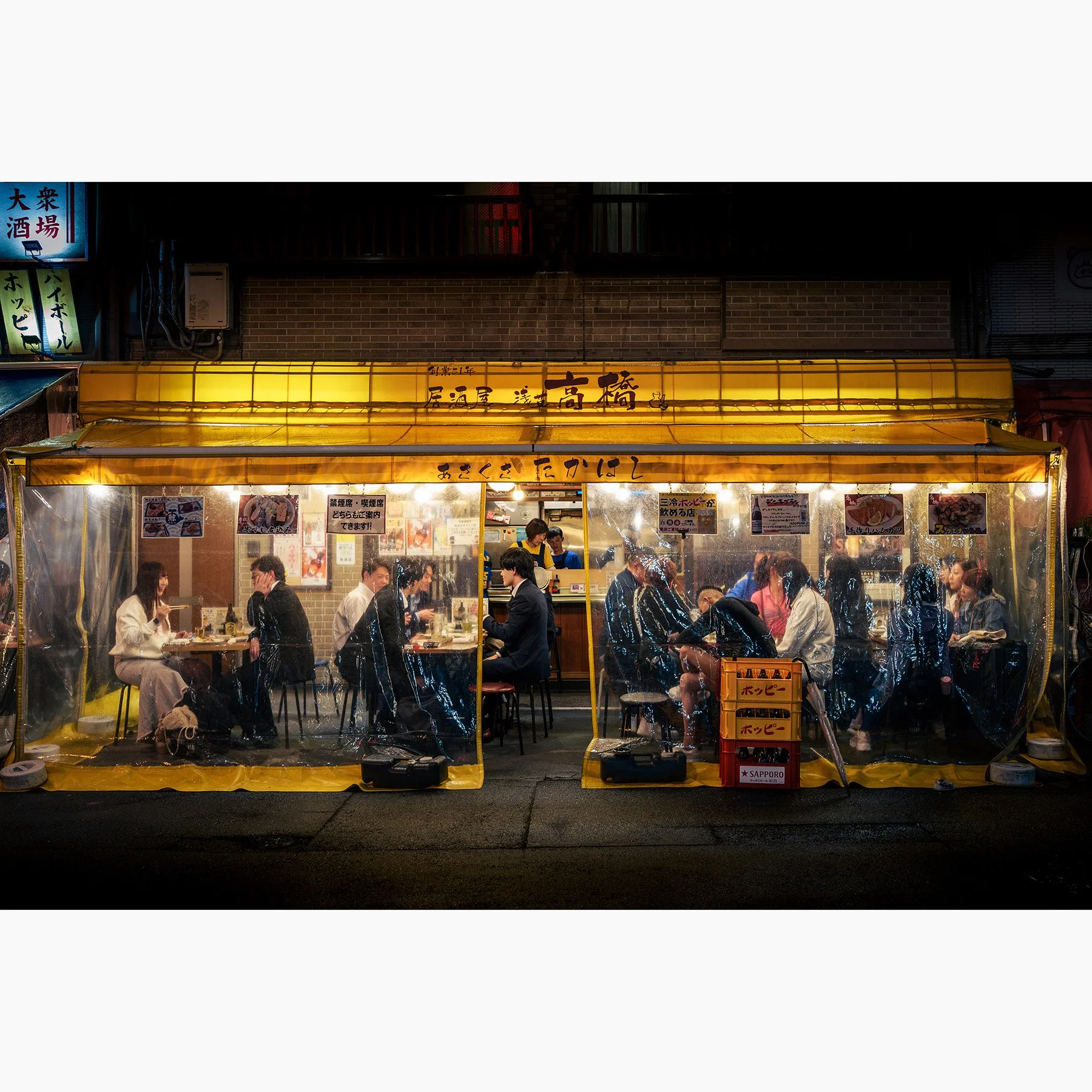 Night street photograph of a busy izakaya in Tokyo's Asakusa district, with diners visible through clear plastic curtains beneath a brightly illuminated yellow awning.