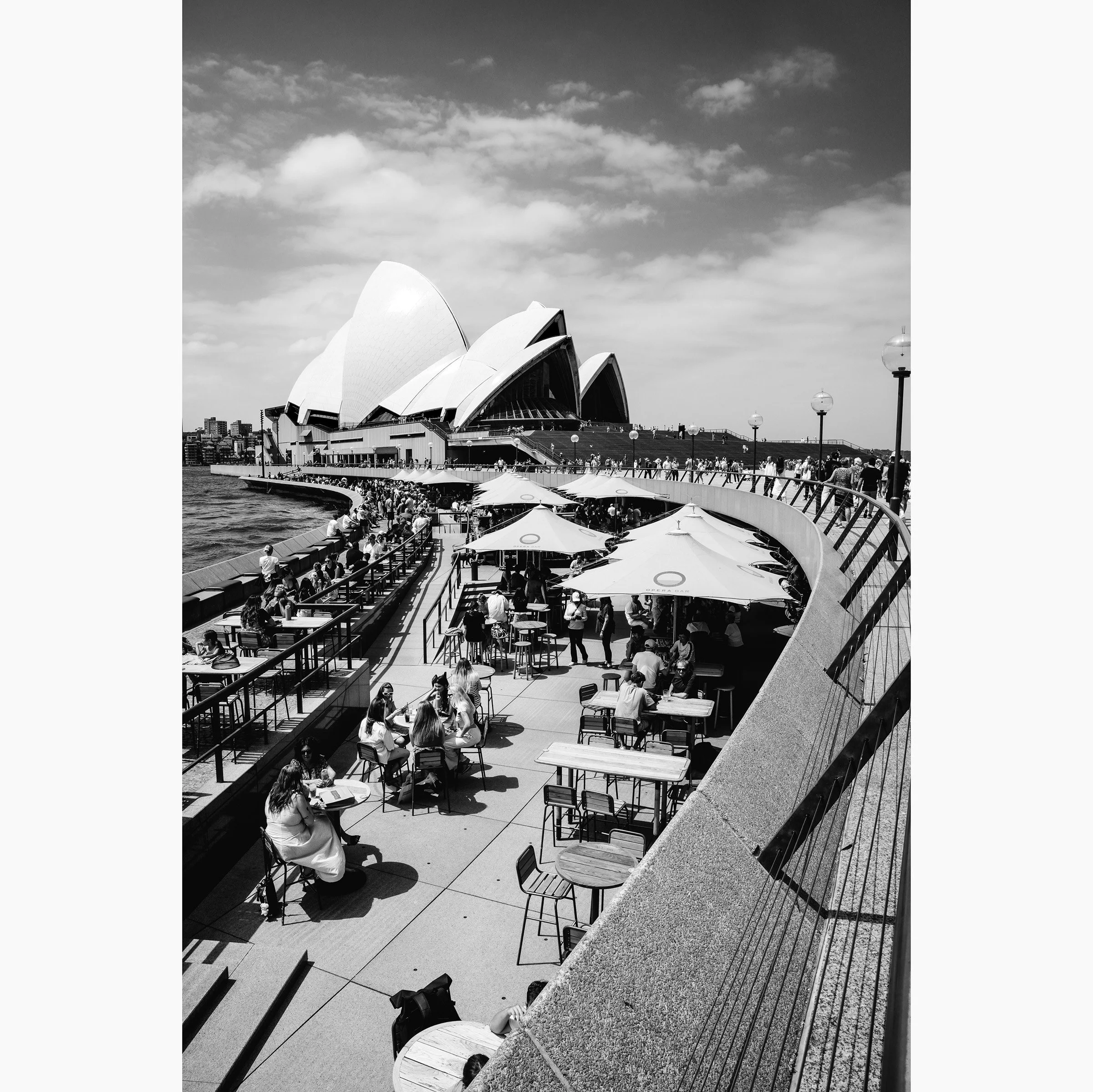 Black and white photograph of the Sydney Opera House and Circular Quay promenade, with waterfront dining, harbour views and crowds gathered along the foreshore.
