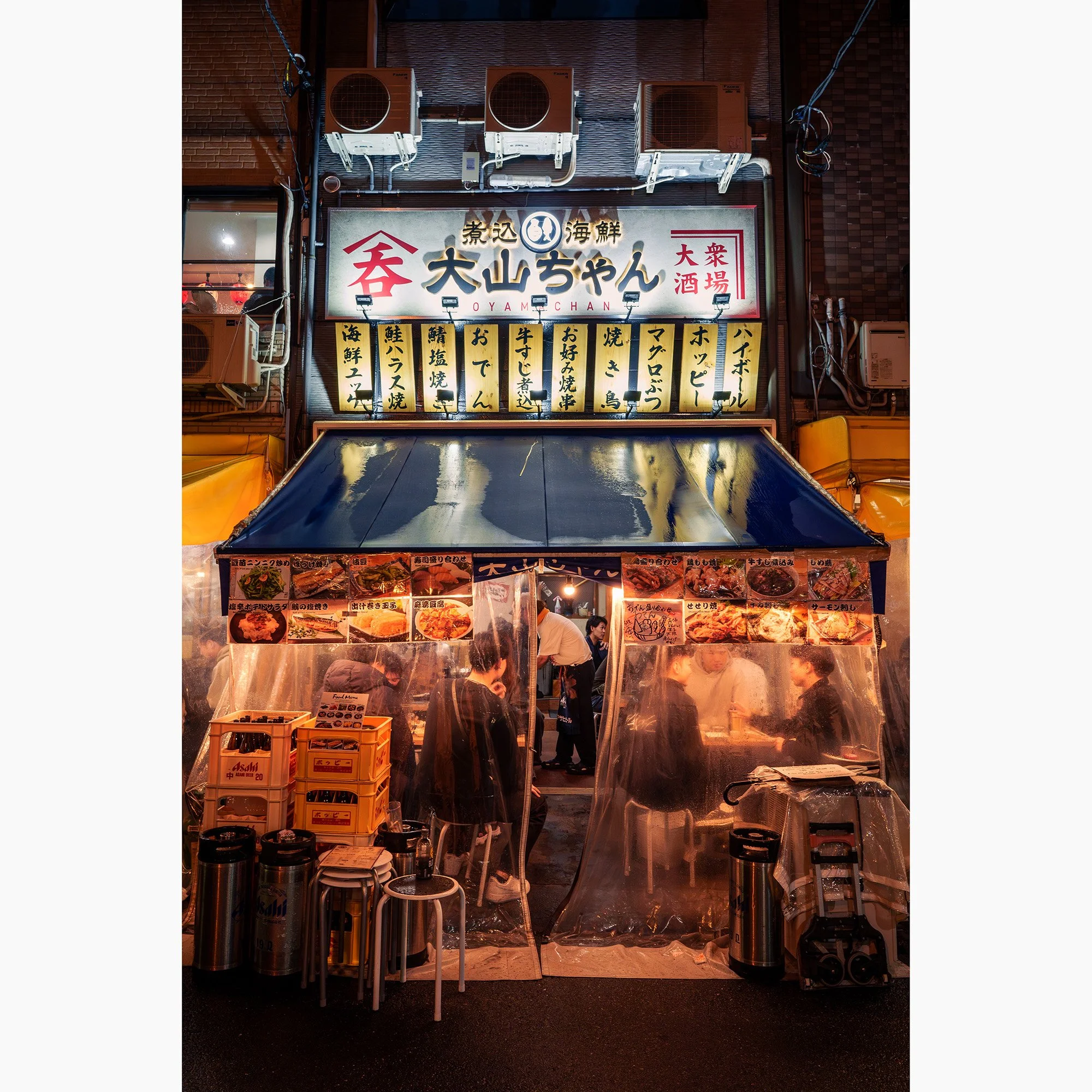Night street photograph of a brightly lit Tokyo izakaya, with Japanese signage, diners and clear plastic curtains around the entrance.