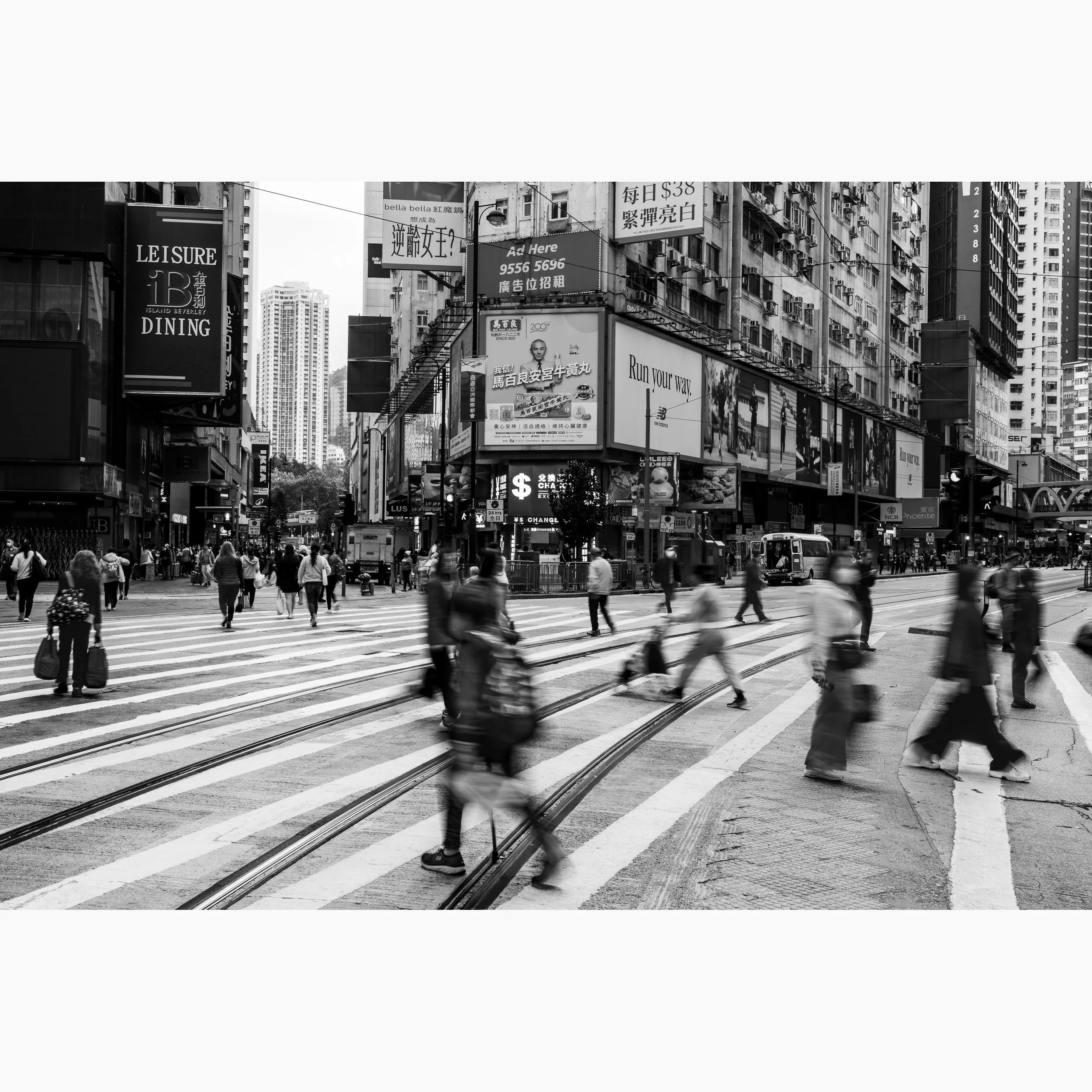 Black and white Hong Kong street photography print featuring blurred pedestrians crossing tram tracks between dense urban buildings and illuminated city signage.