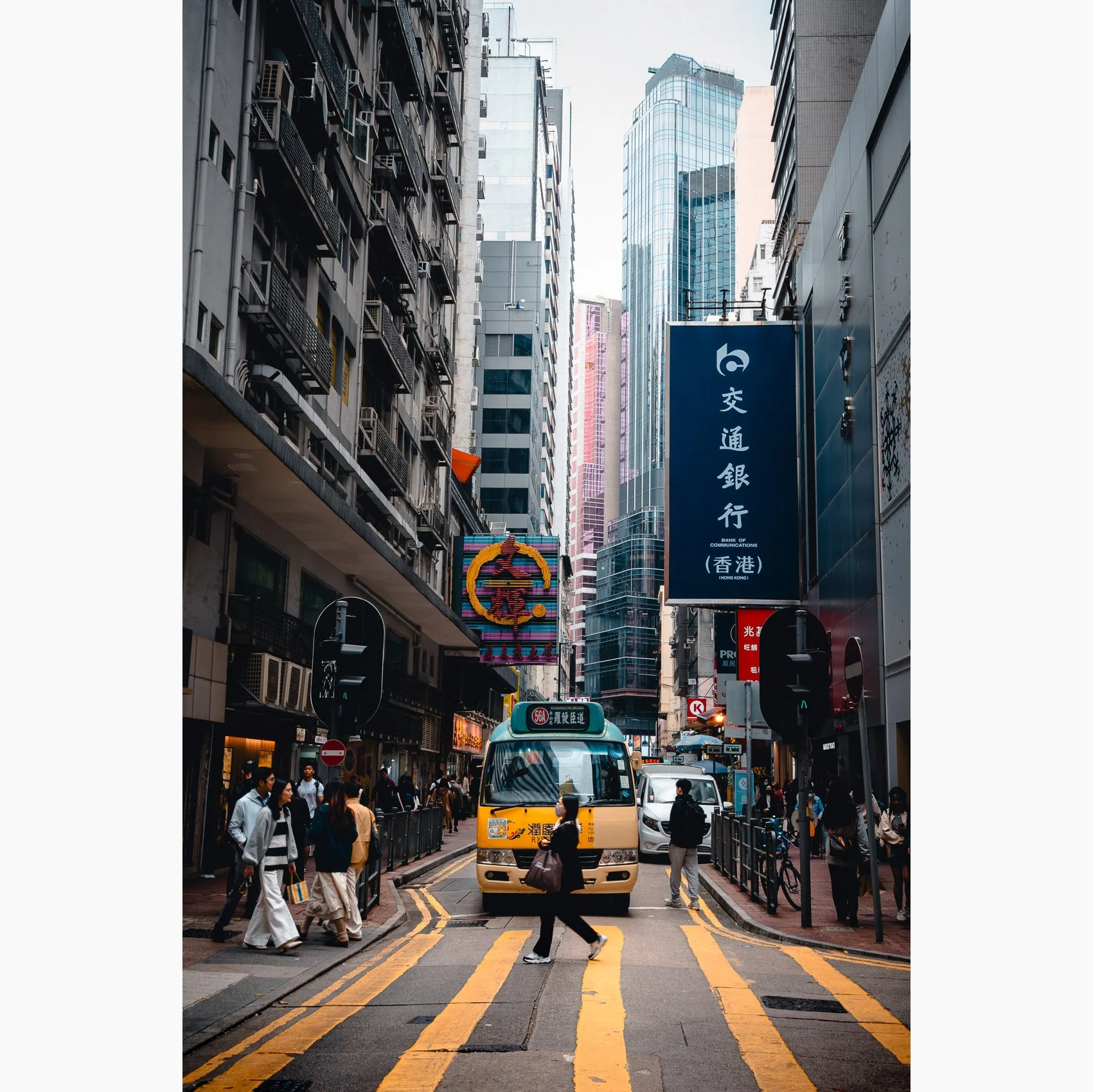 Hong Kong street photography print featuring a yellow minibus travelling through a dense urban street scene with pedestrians, signage and tall city buildings.