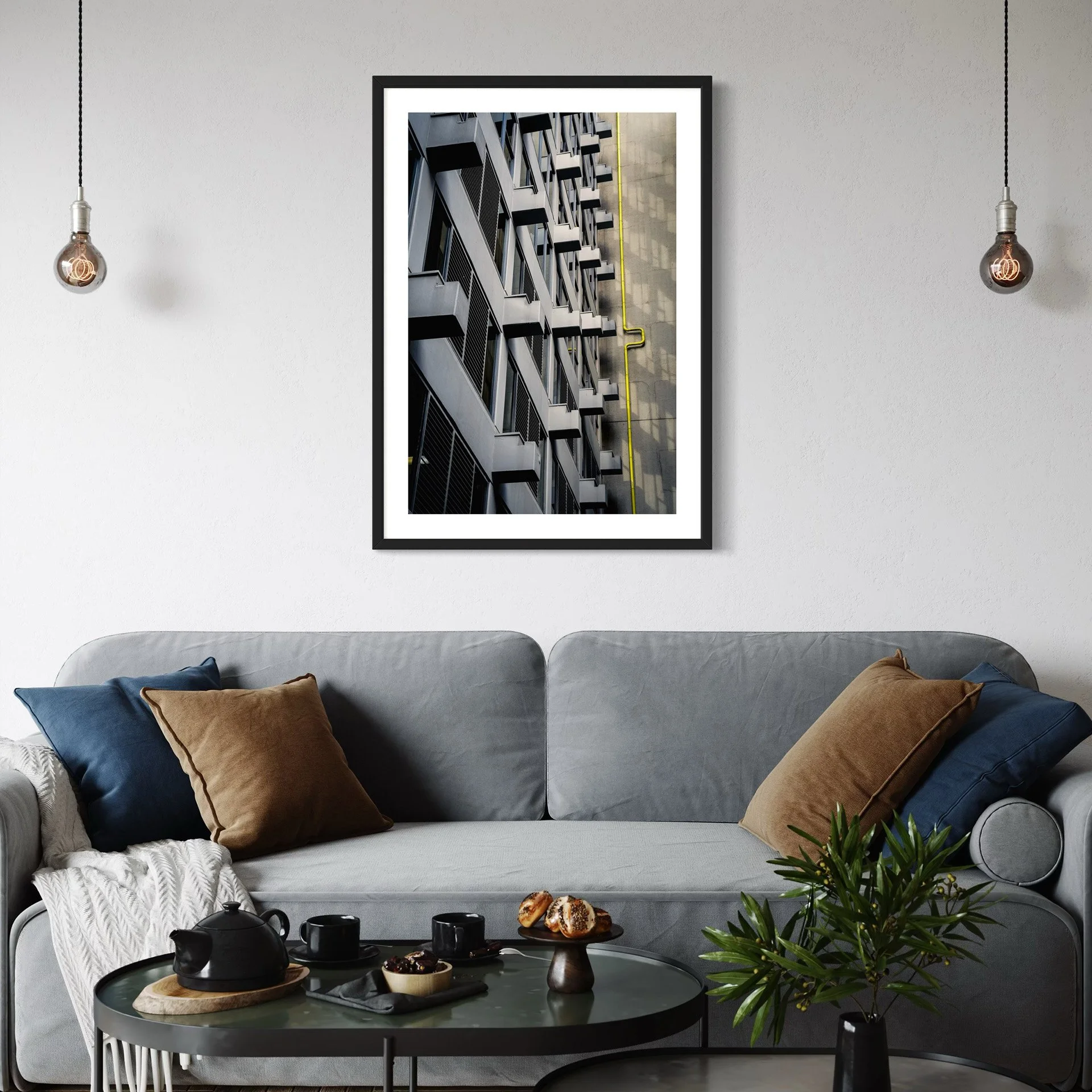 Modern lounge with a framed urban architecture print showing geometric balconies and a yellow pipe displayed above a grey sofa.
