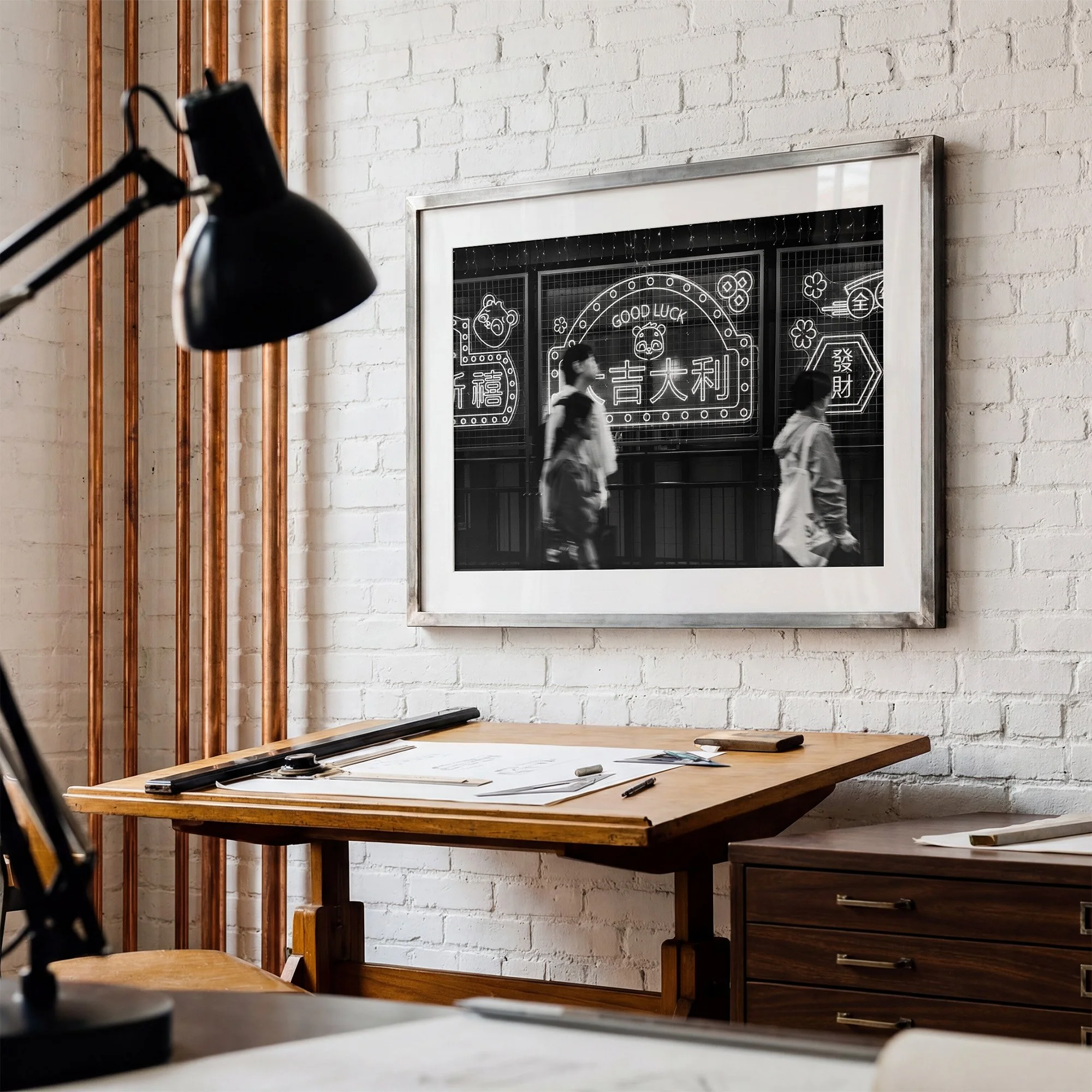 Hong Kong black and white street photography print featuring illuminated good luck signs and passing pedestrians, displayed in a silver frame on a white brick wall.