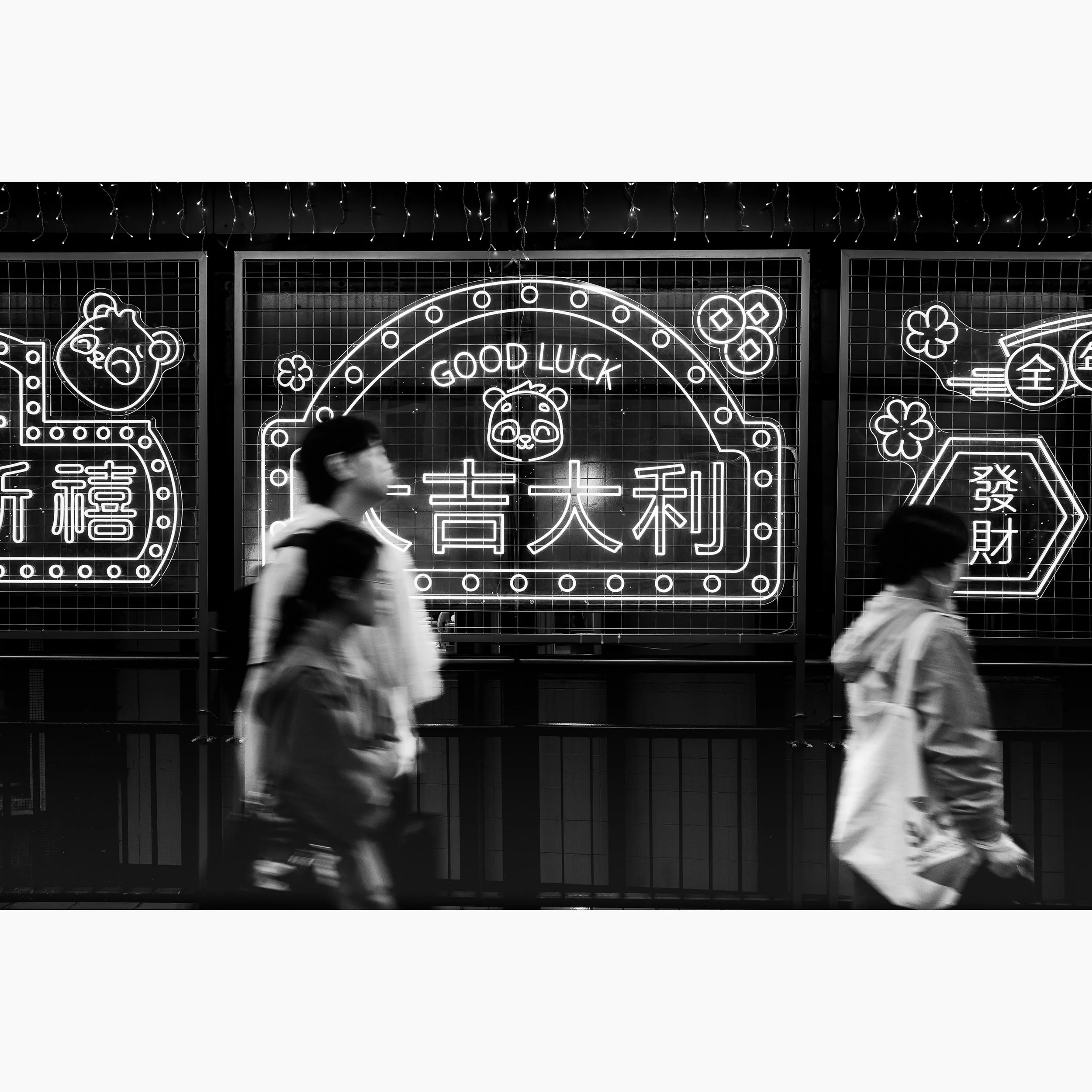 Black and white street photograph of pedestrians walking past glowing neon “Good Luck” signage in Hong Kong at night.